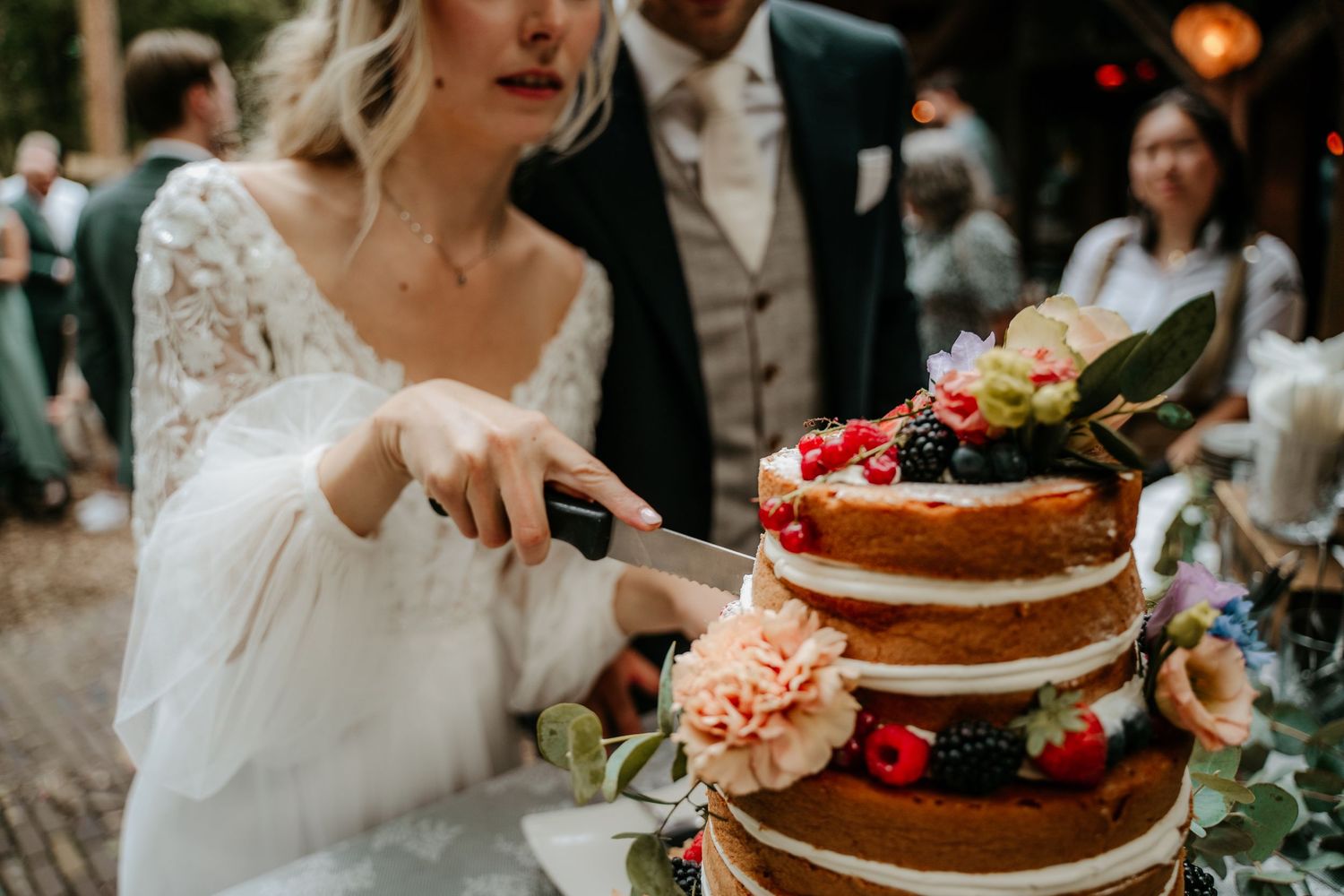 Wedding couple cutting into naked cake decorated with fresh berries and flowers.