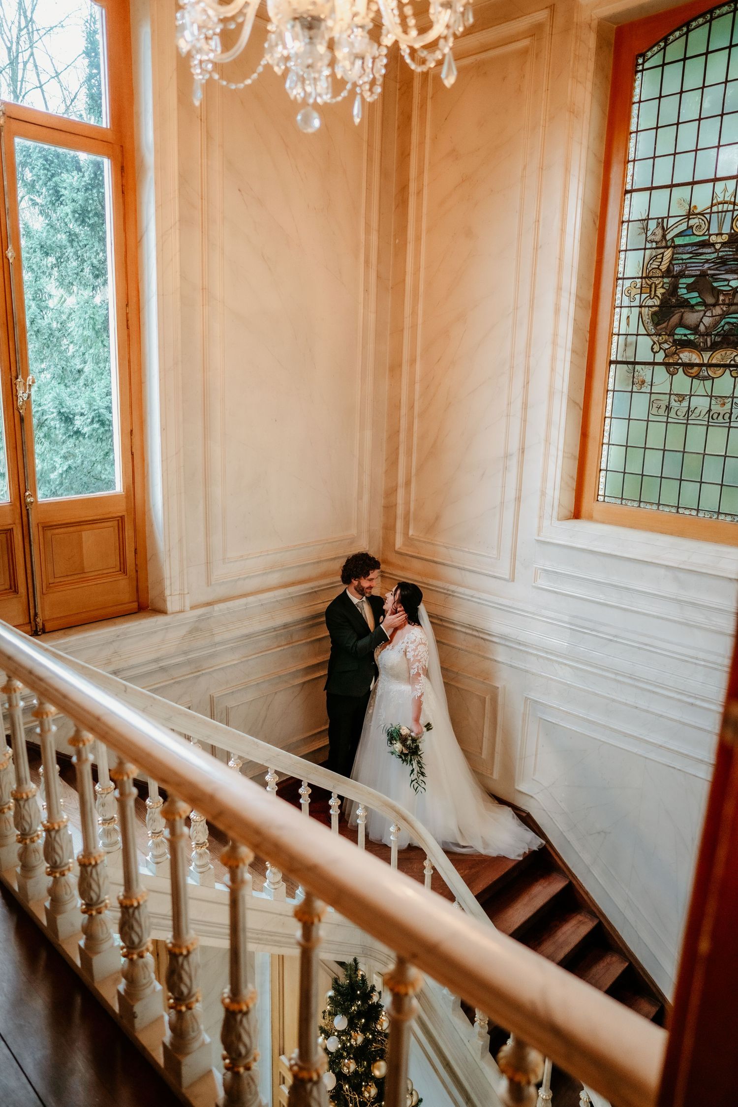 Newlyweds share intimate moment on elegant staircase under crystal chandelier.