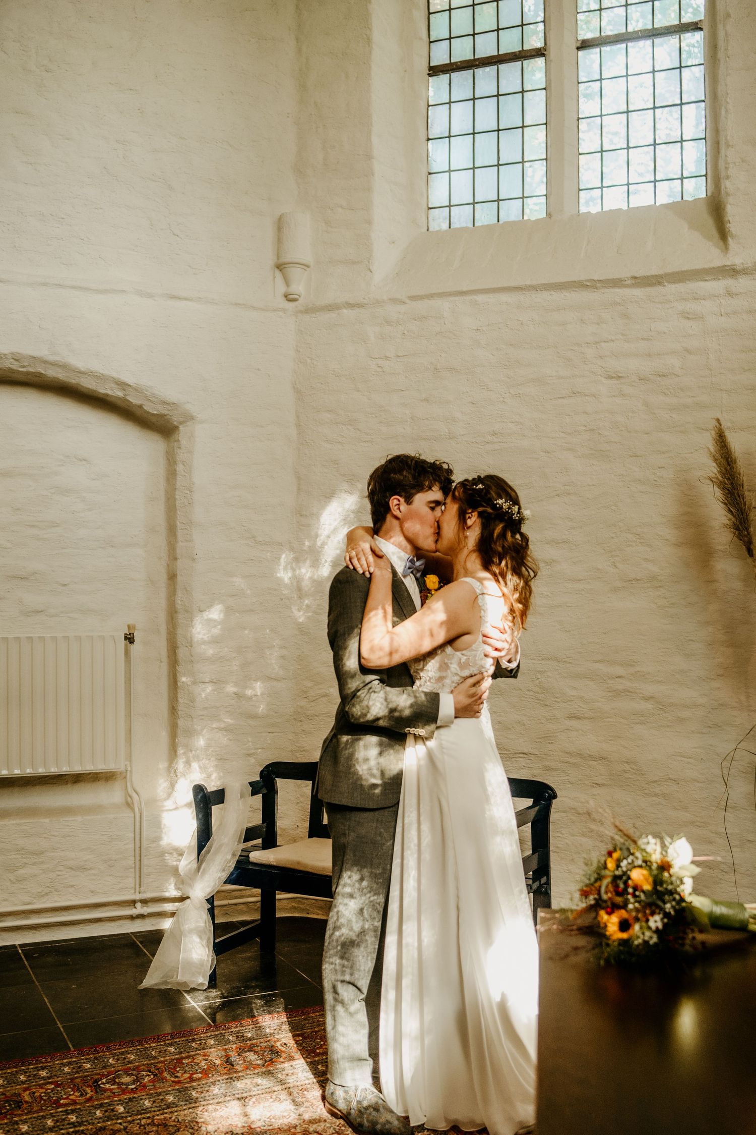 A romantic embrace between newlyweds in a rustic church interior with sunlit windows and white walls.
