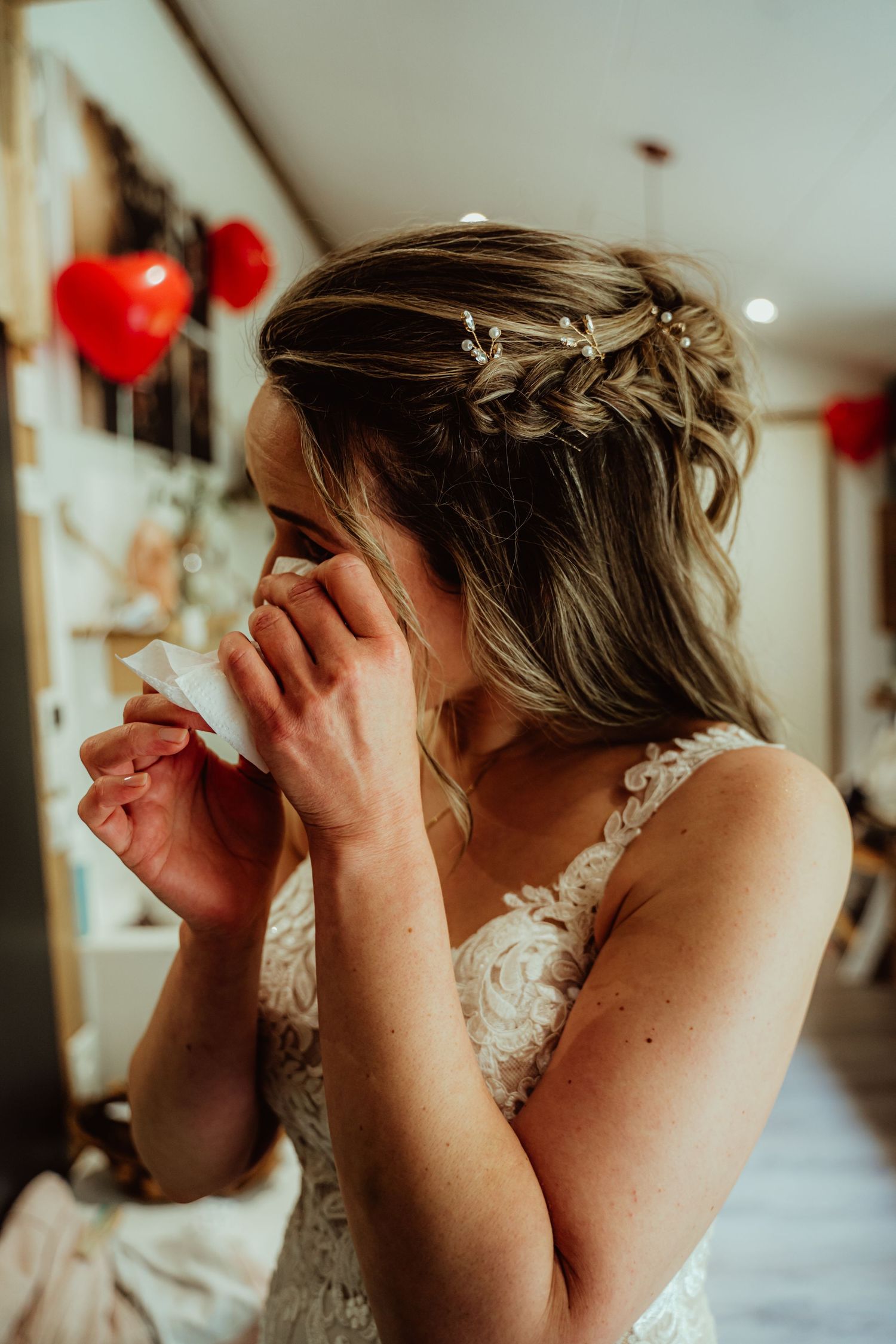 Emotional bride in lace dress with braided hair detail and red heart decorations in background.