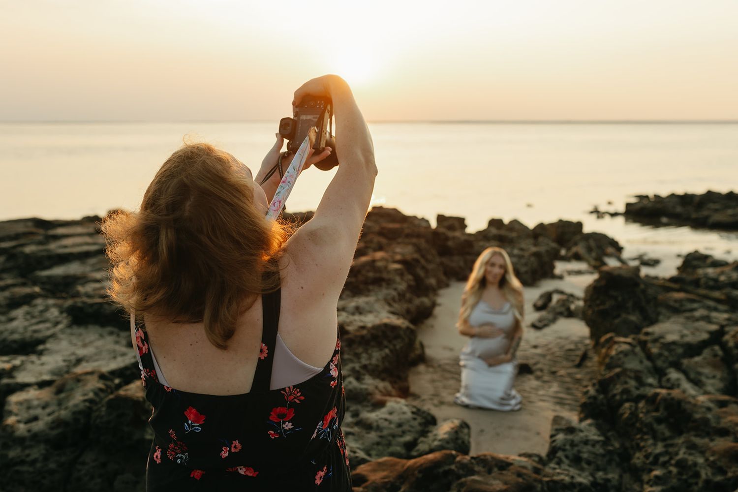 Photographer capturing sunset beach photos on rocky coastline during golden hour.