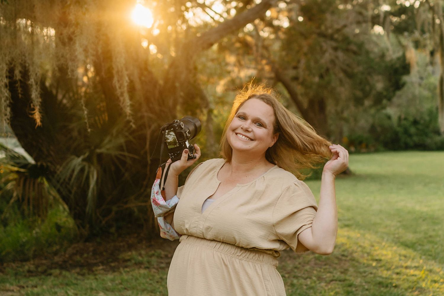 Jessica Life, your Jacksonville newborn photographer in a beige dress holding camera and twirling hair in golden sunset light.