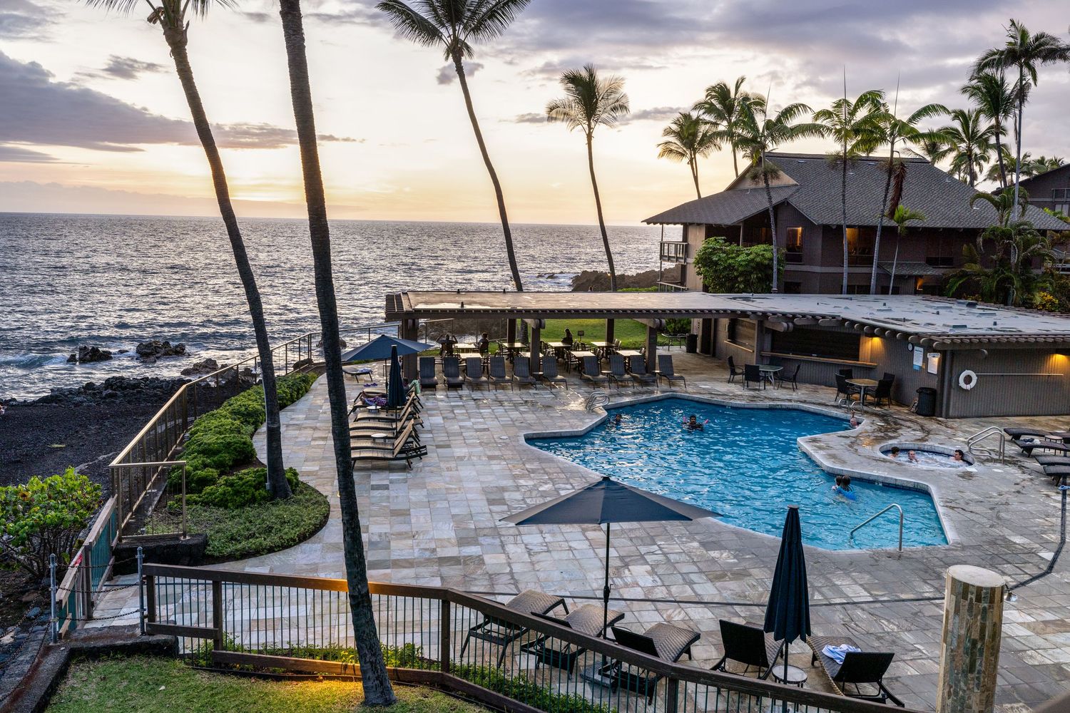 Beachfront resort with swimming pool overlooking palm trees and ocean at sunset.