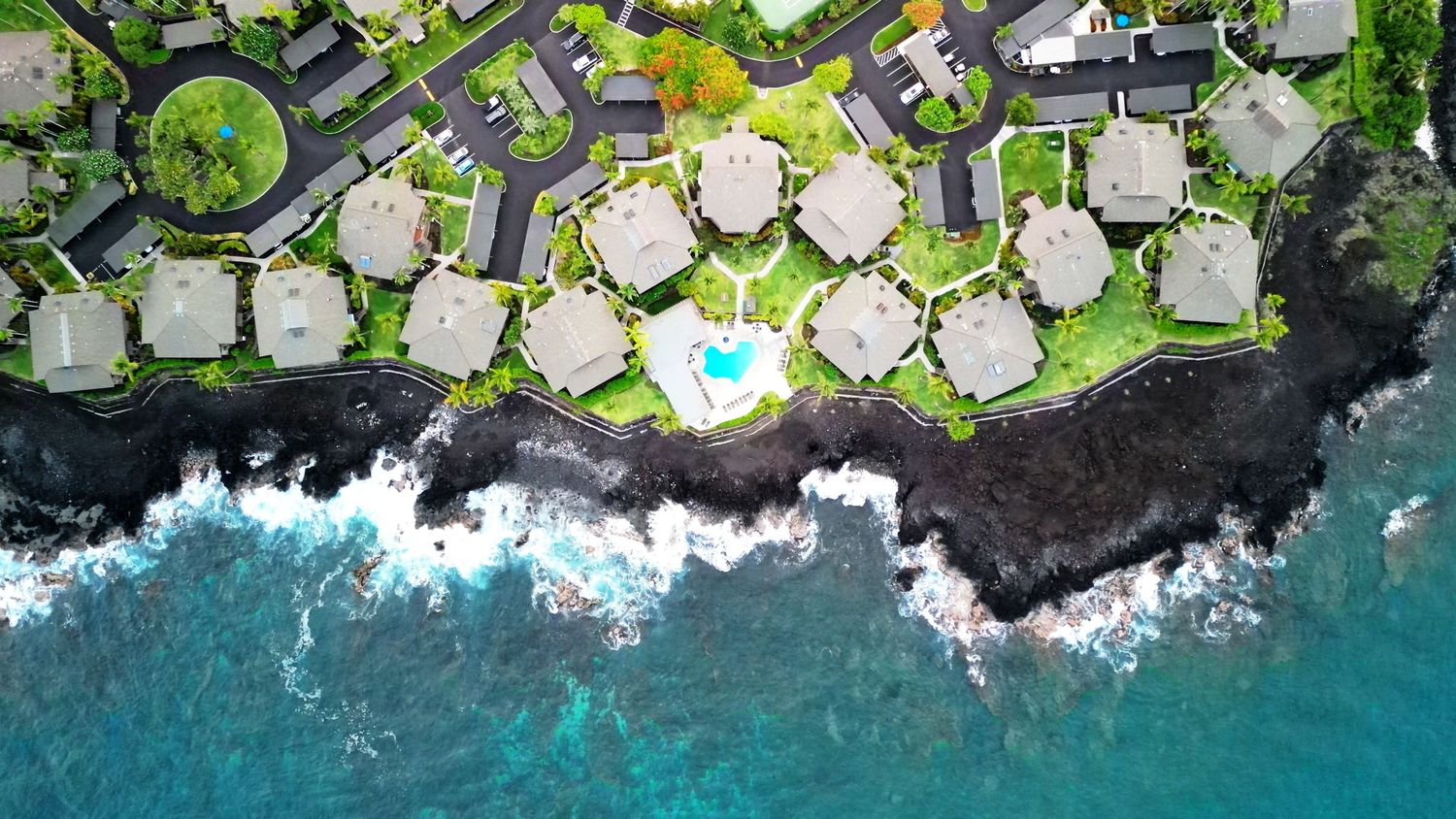 Birds eye view of oceanfront vacation homes with pool and black lava rock coastline.