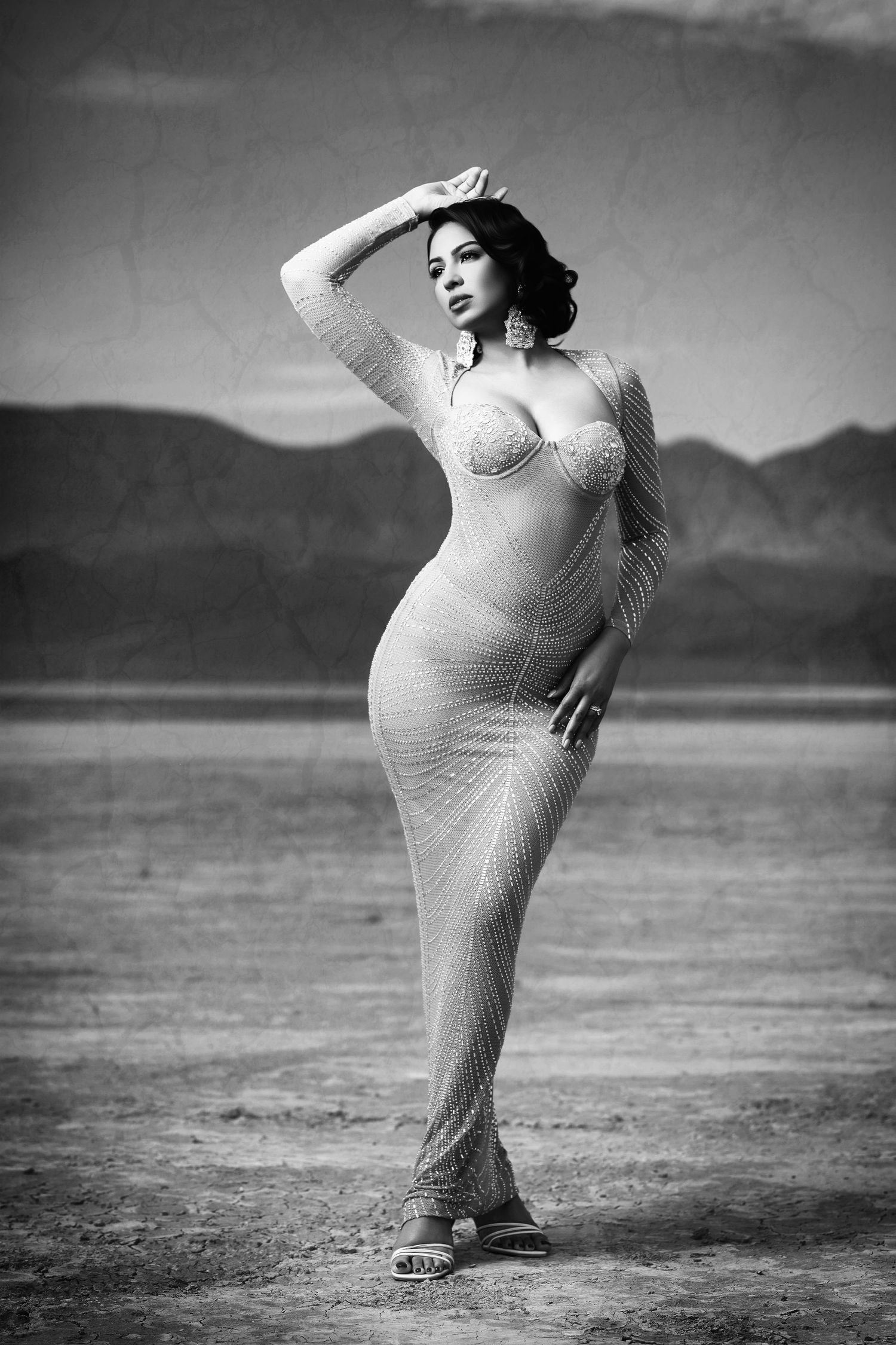 A dramatic black and white desert portrait of someone in a form-fitting dress posing against mountain backdrop.