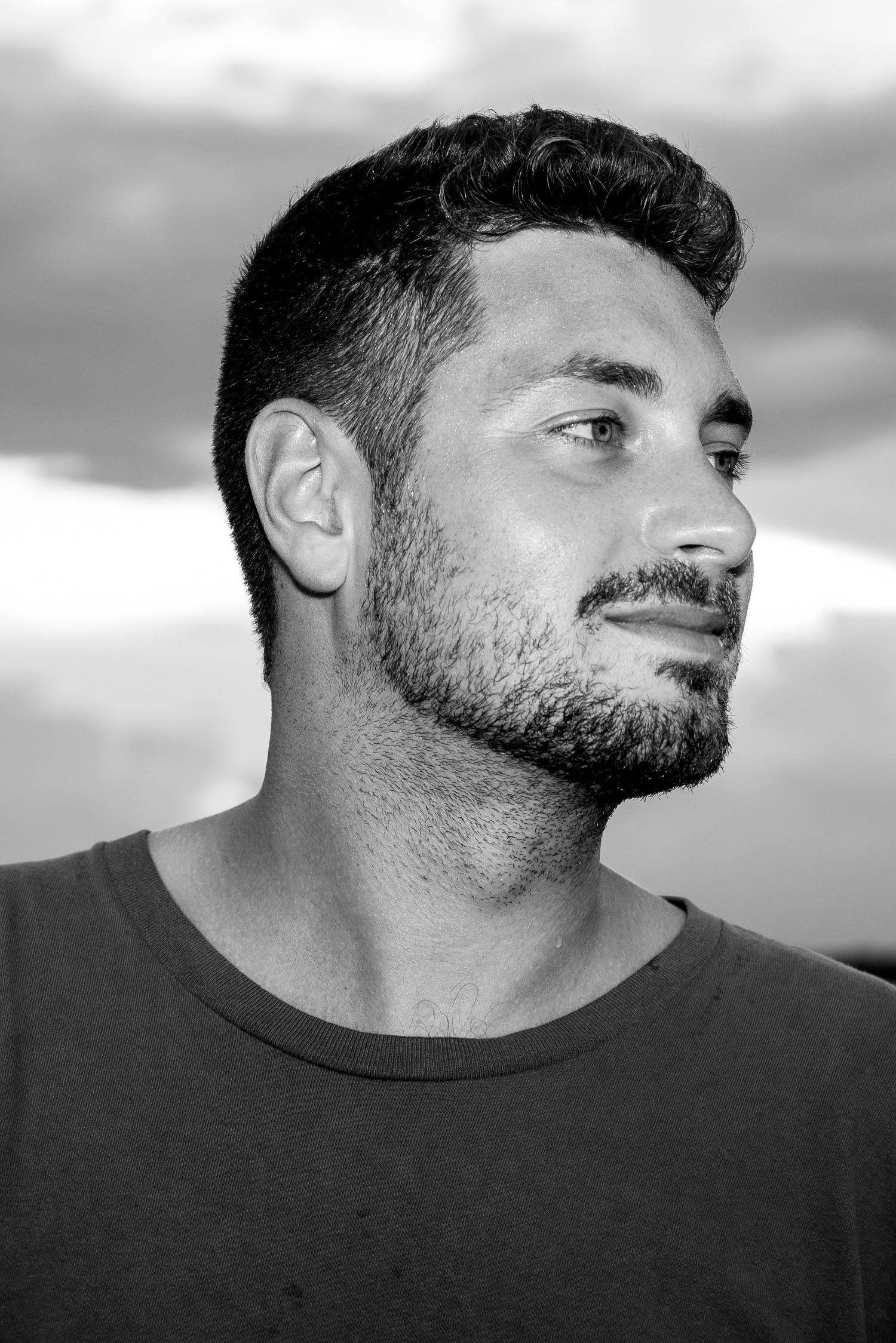 Black and white profile portrait of person with short dark hair and beard against cloudy sky.