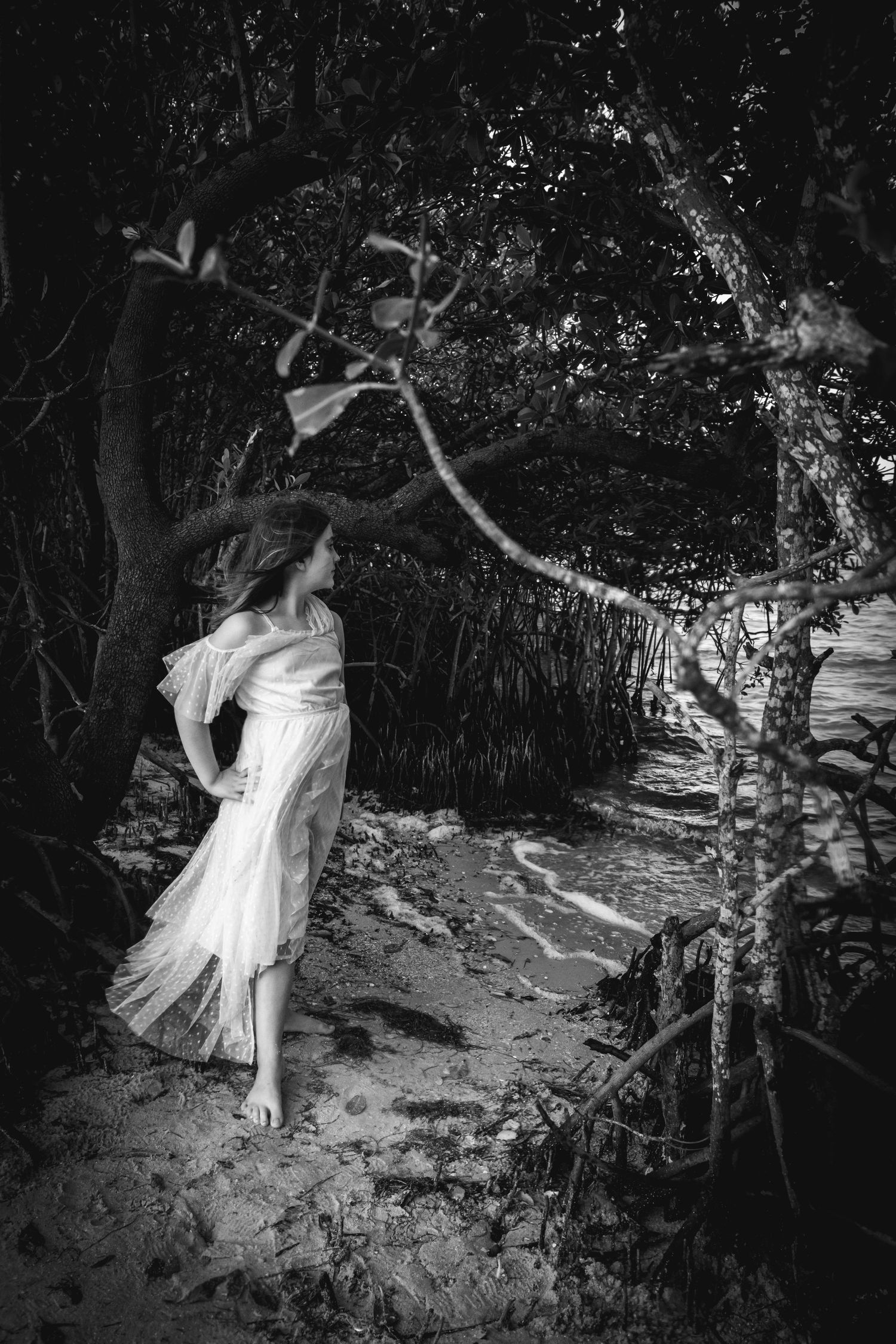 Ethereal black and white photo of a figure in flowing white dress walking along a wooded path.