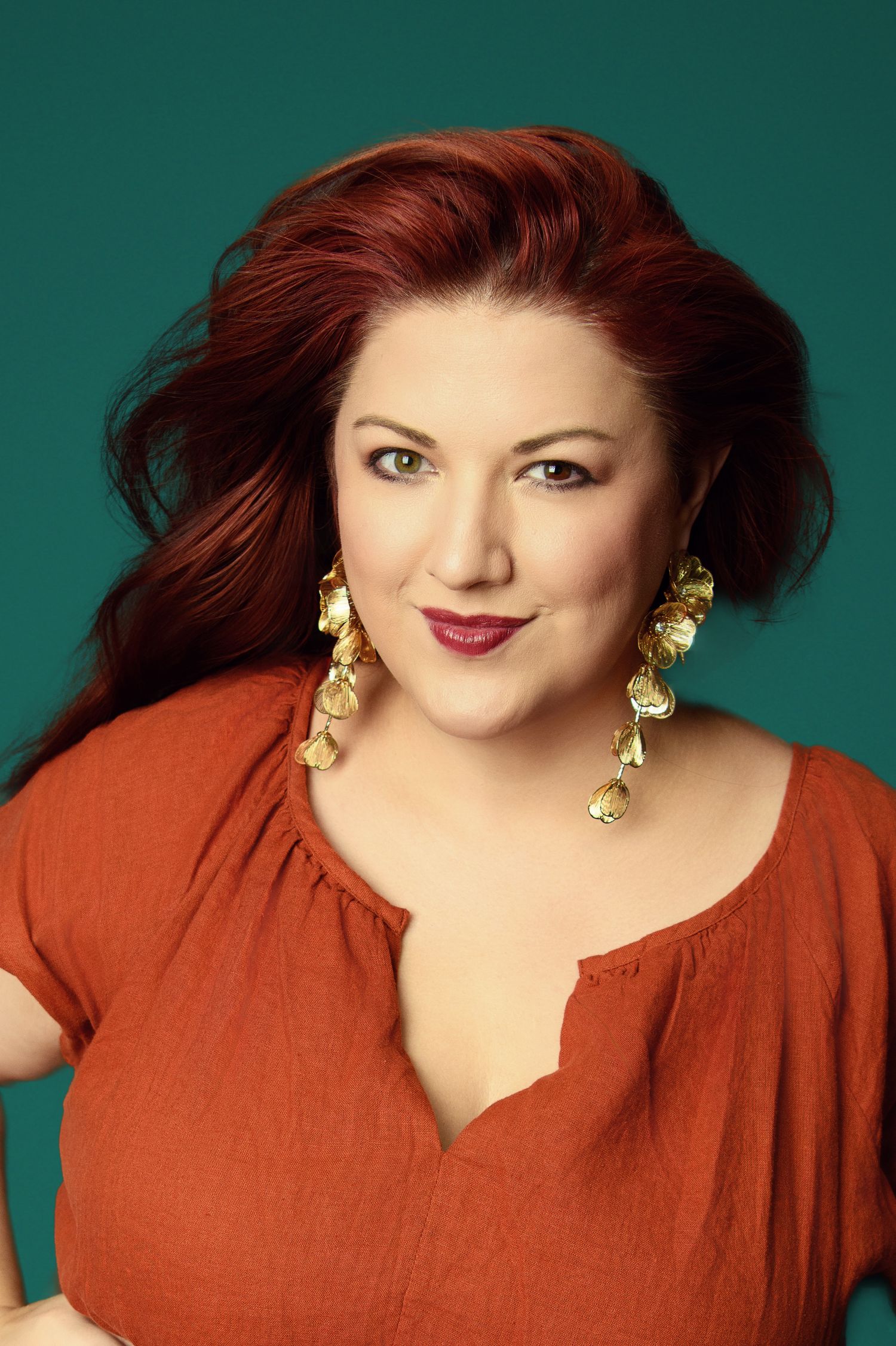 Professional studio portrait with teal background featuring warm rusty orange tones and gold jewelry.