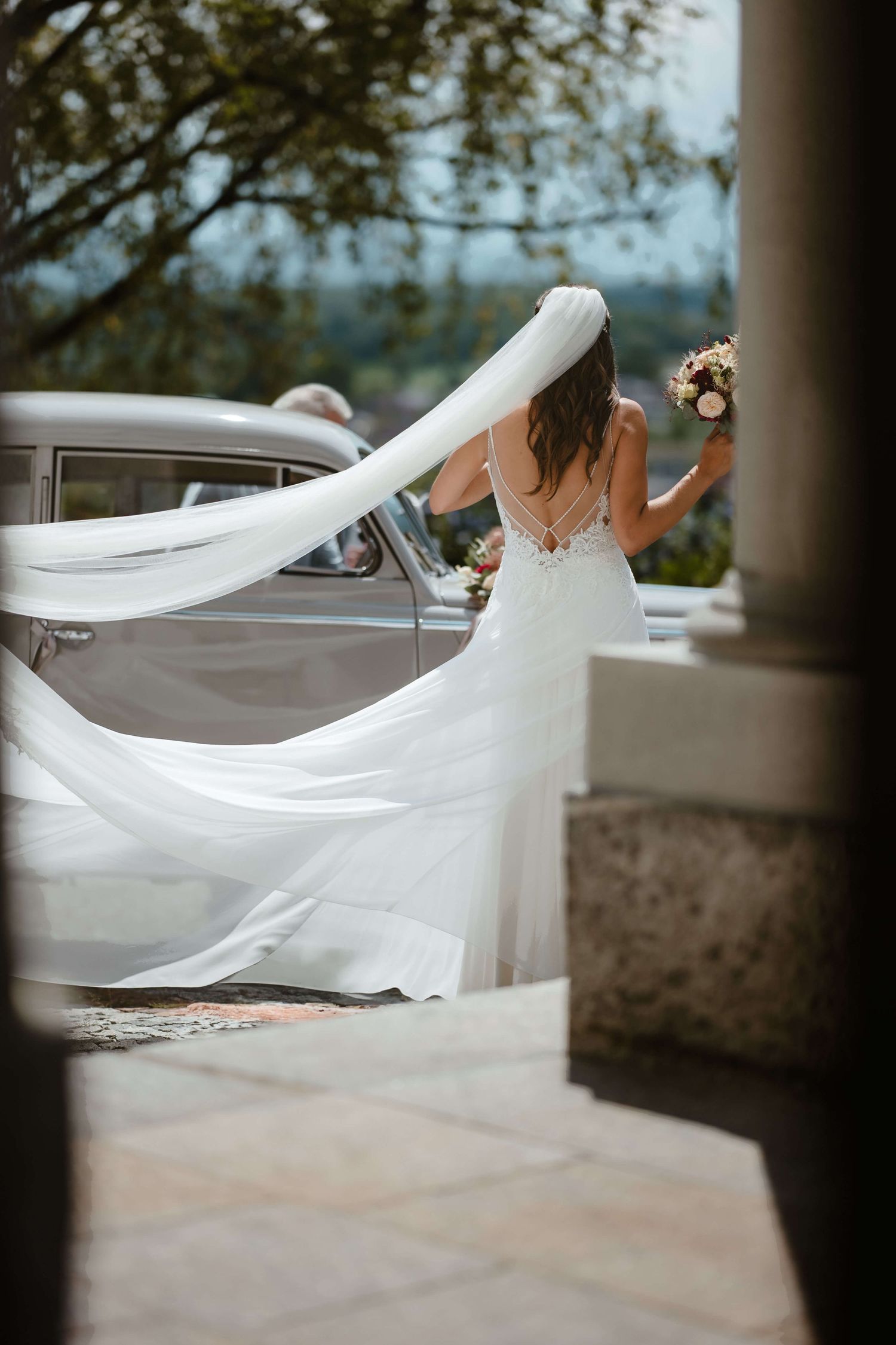 Simone vor dem Hochzeitsauto mit wehendem Schleier – stilvoller Moment in Vorarlberg, fotografiert von NuMa Fotografie