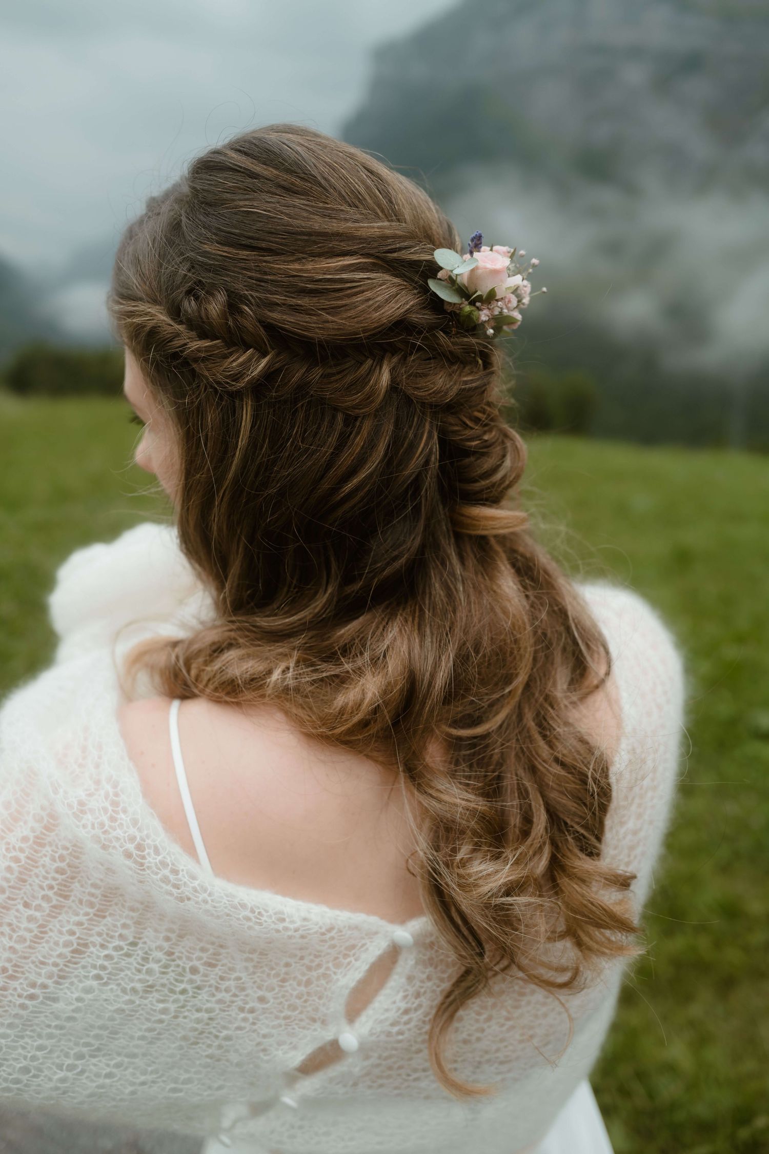 Die Braut steht in einem weißen Kleid auf einer Wiese in Vorarlberg, ihre detailverliebte Hochsteckfrisur mit Blumen wird von hinten gezeigt