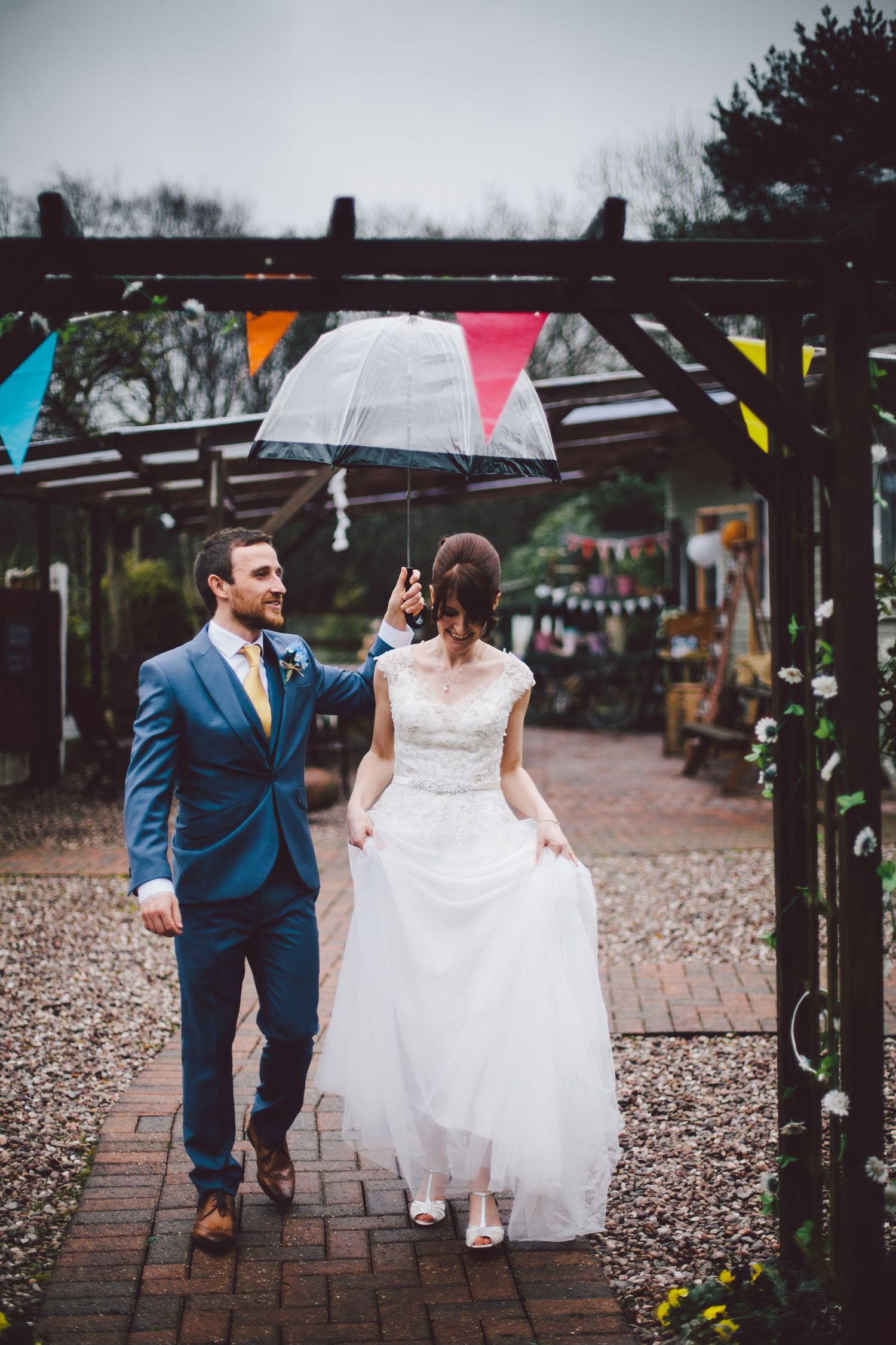 A couple shares an umbrella while walking under a decorated pergola with colorful bunting at a rustic outdoor wedding venue.