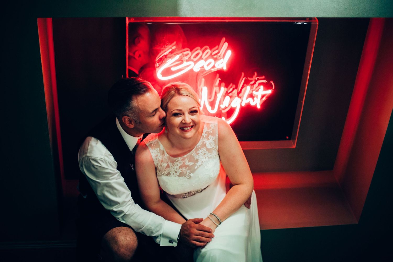 A couple sits together beneath a glowing red neon sign in a dimly lit space, creating a romantic mood.