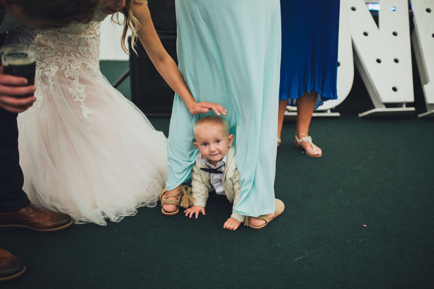 A playful toddler crawls between formal wedding attire on a green carpet at a reception.