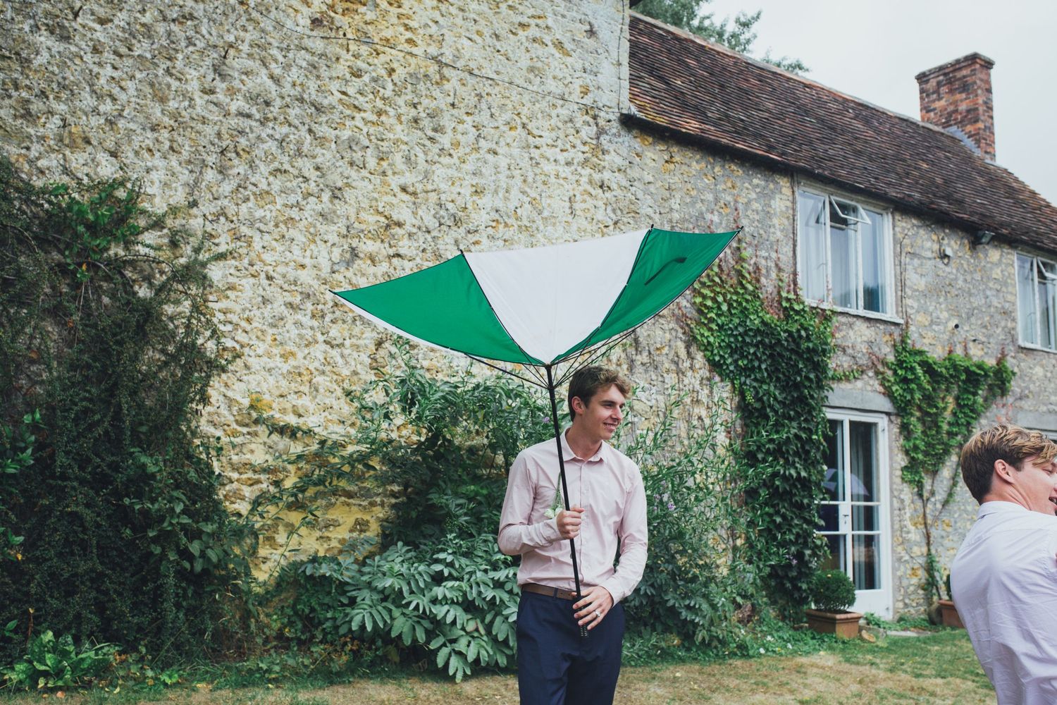 A green umbrella shields someone at a rustic Somerset country house with stone walls and traditional windows.