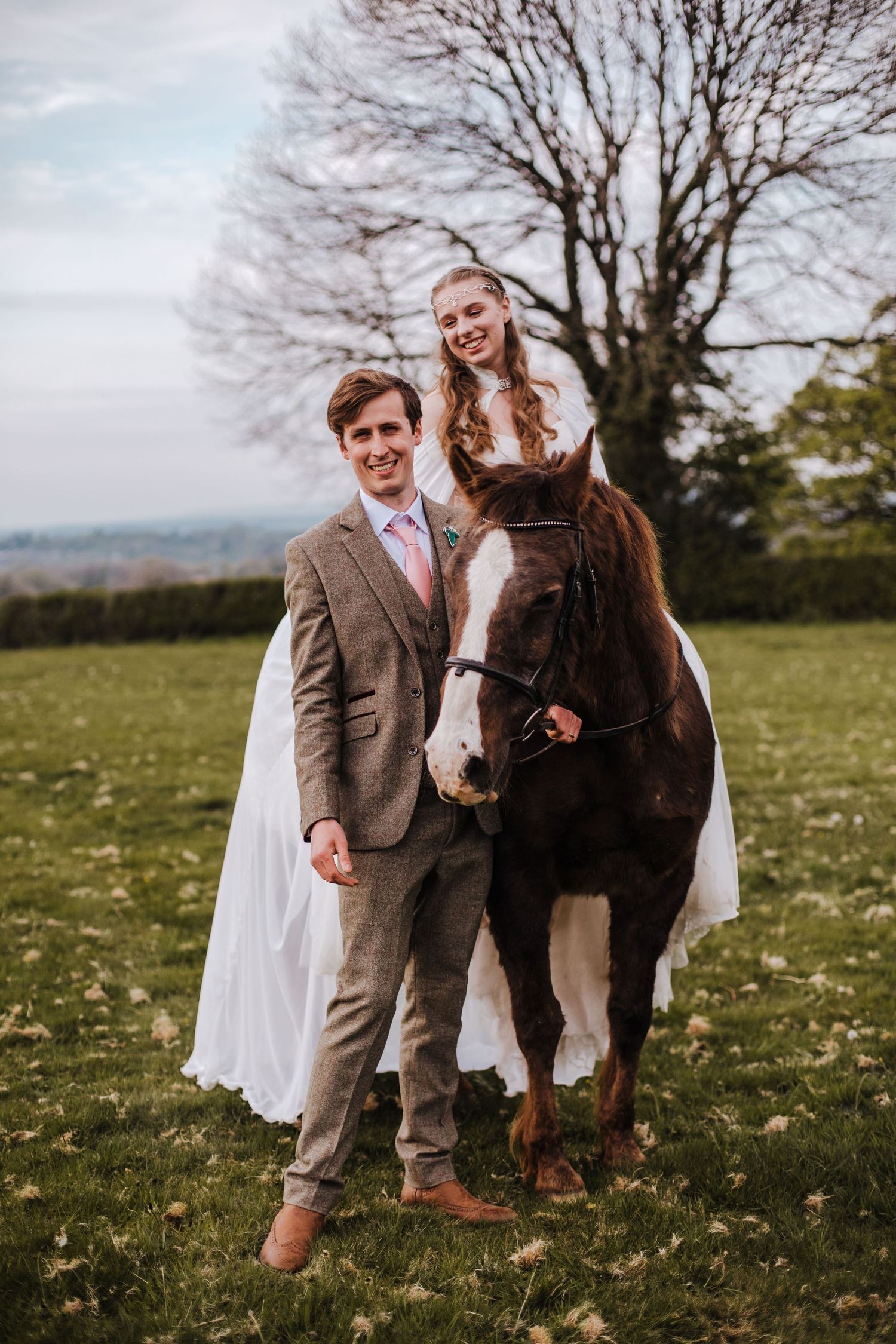 Rustic wedding portrait featuring a couple in formal attire posing with their brown and white horse in a scenic field.