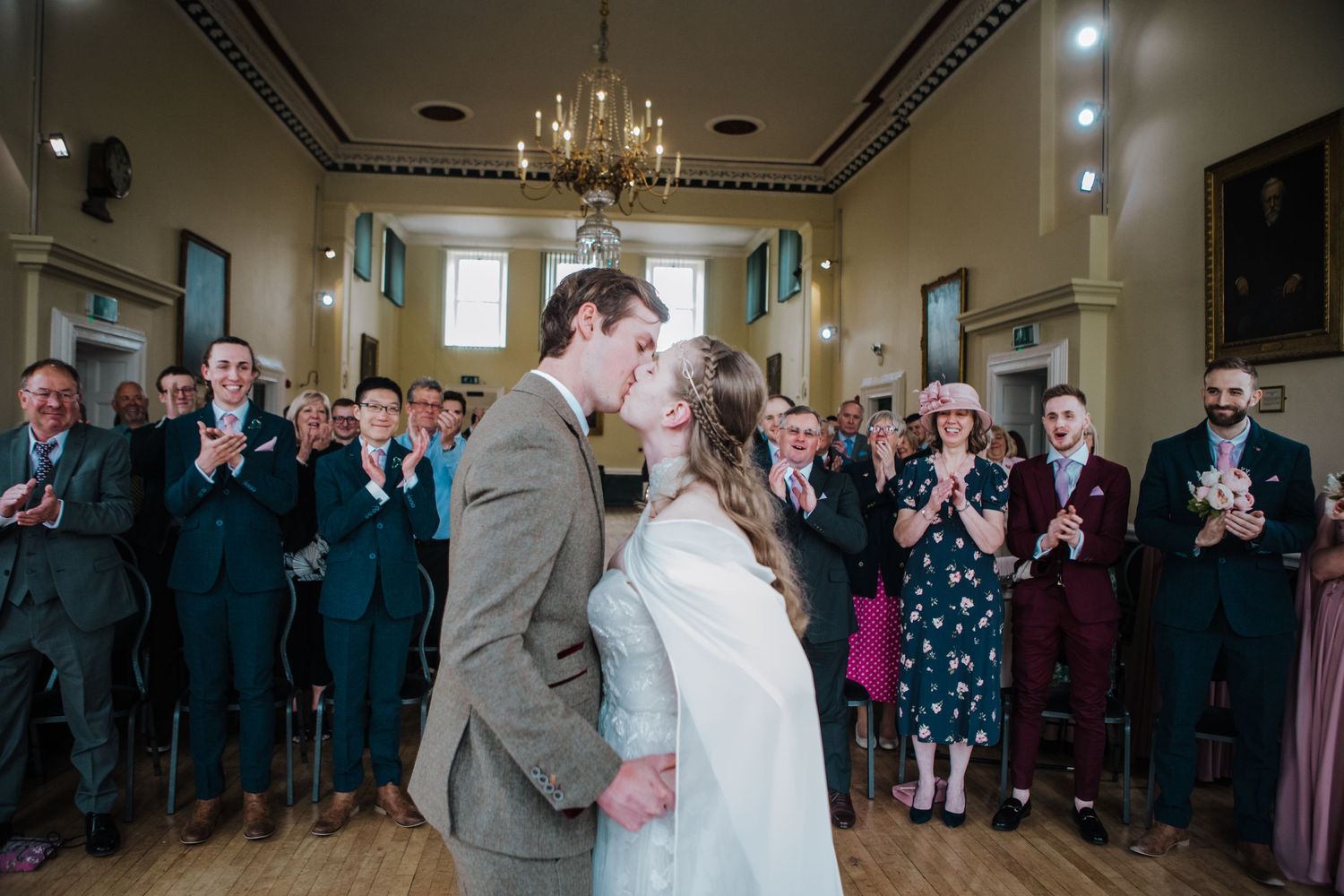 A couple shares their first kiss at their wedding ceremony in an elegant ballroom while guests applaud.