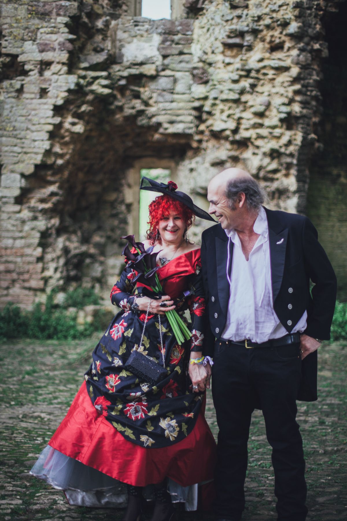 Gothic, Victorian-style couple in formal attire share a tender moment against weathered stone castle wall ruins.
