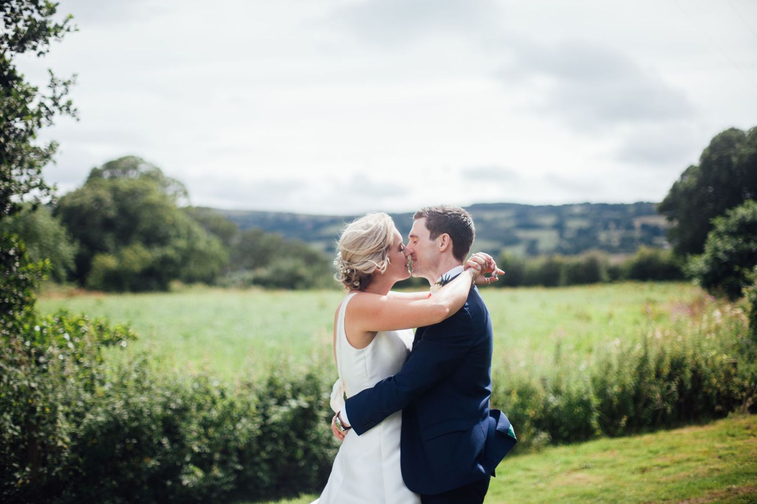 Somerset wedding photographer -A couple in wedding attire embrace in a romantic kiss against a scenic countryside landscape on a summer day.