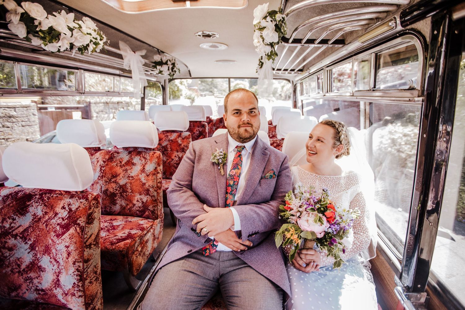 A joyful scene inside a vintage bus with floral seats and a colourful bouquet on a sunny wedding day.