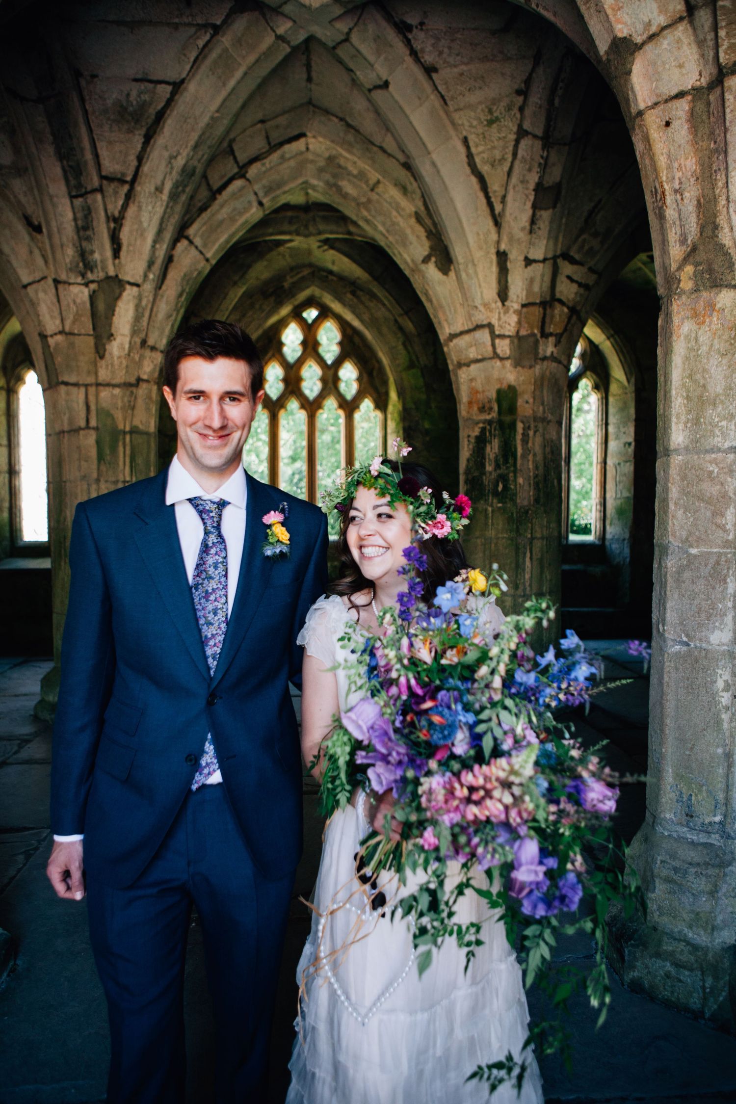 Wedding couple poses in stone archways of historic abbey with stunning purple and pastel floral bouquet.