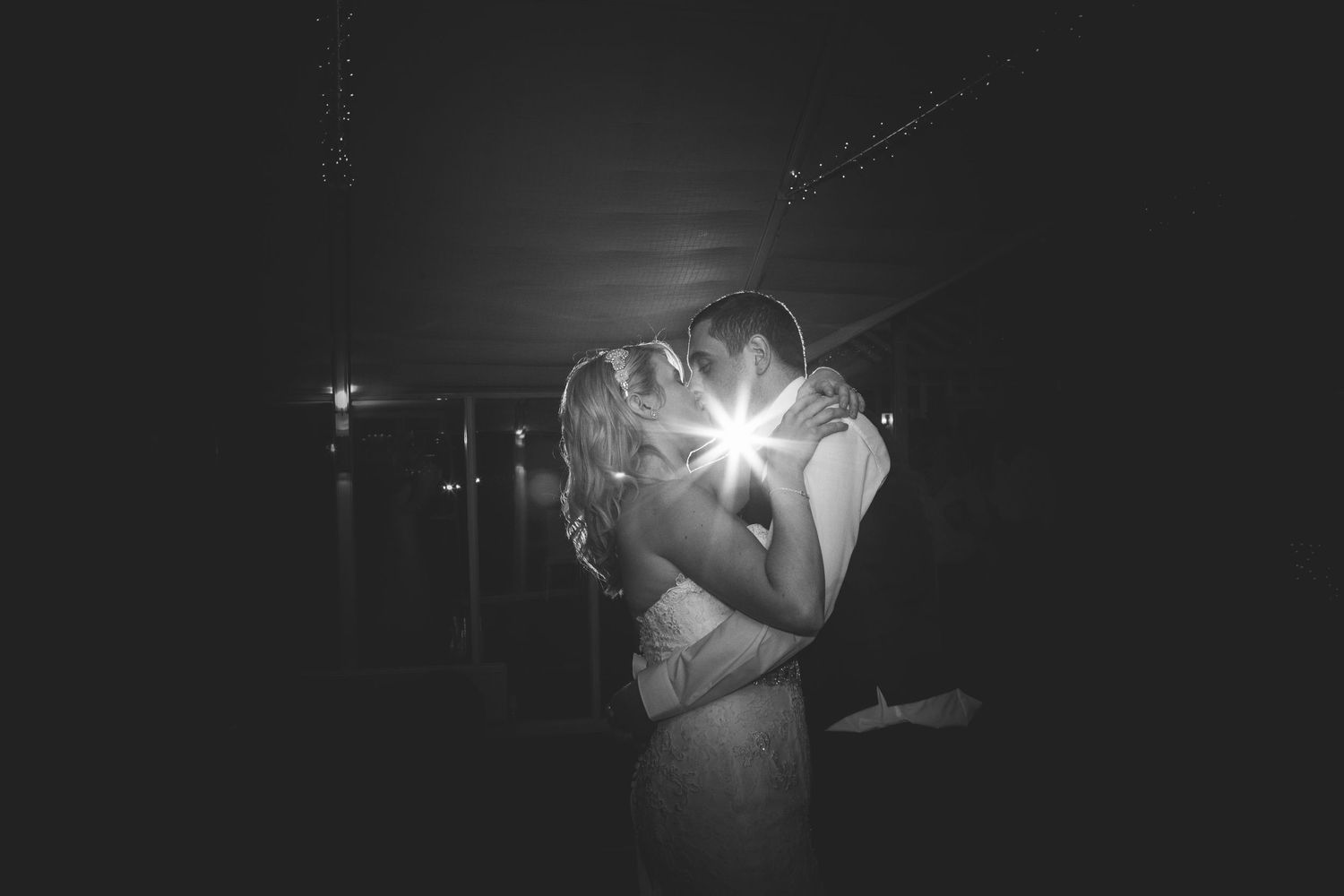 A dramatic black and white wedding photo showing a couple embracing in silhouette with a bright lens flare between them.