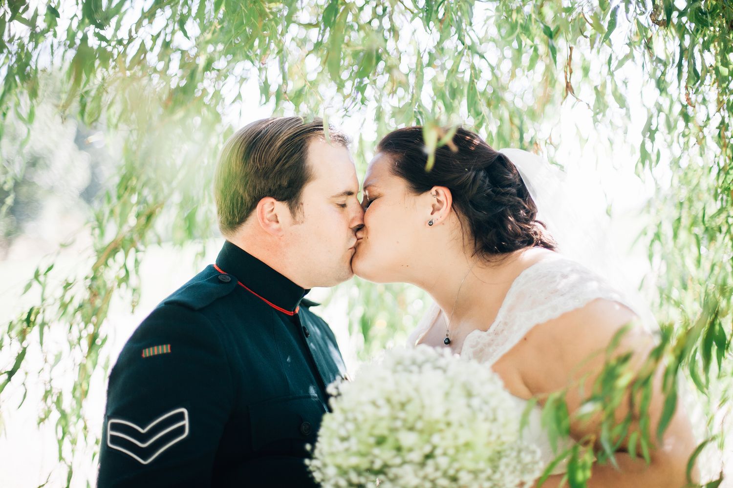 A romantic couple shares a tender kiss under cascading willow branches with white blossoms in soft sunlight.