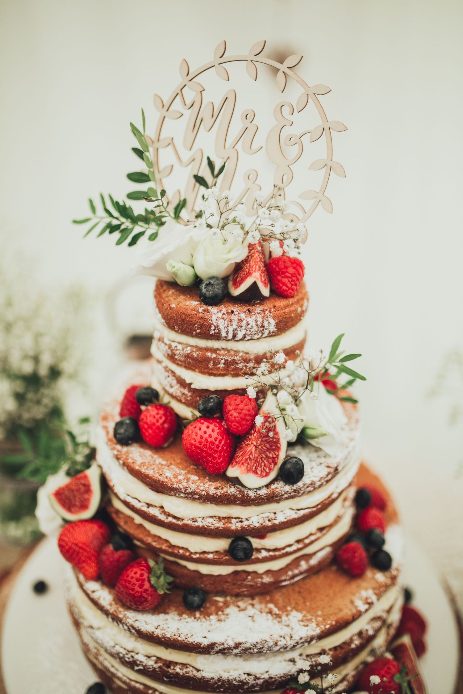 A rustic naked wedding cake decorated with fresh strawberries, figs, and greenery topped with a delicate Mr & Mrs cake topper.