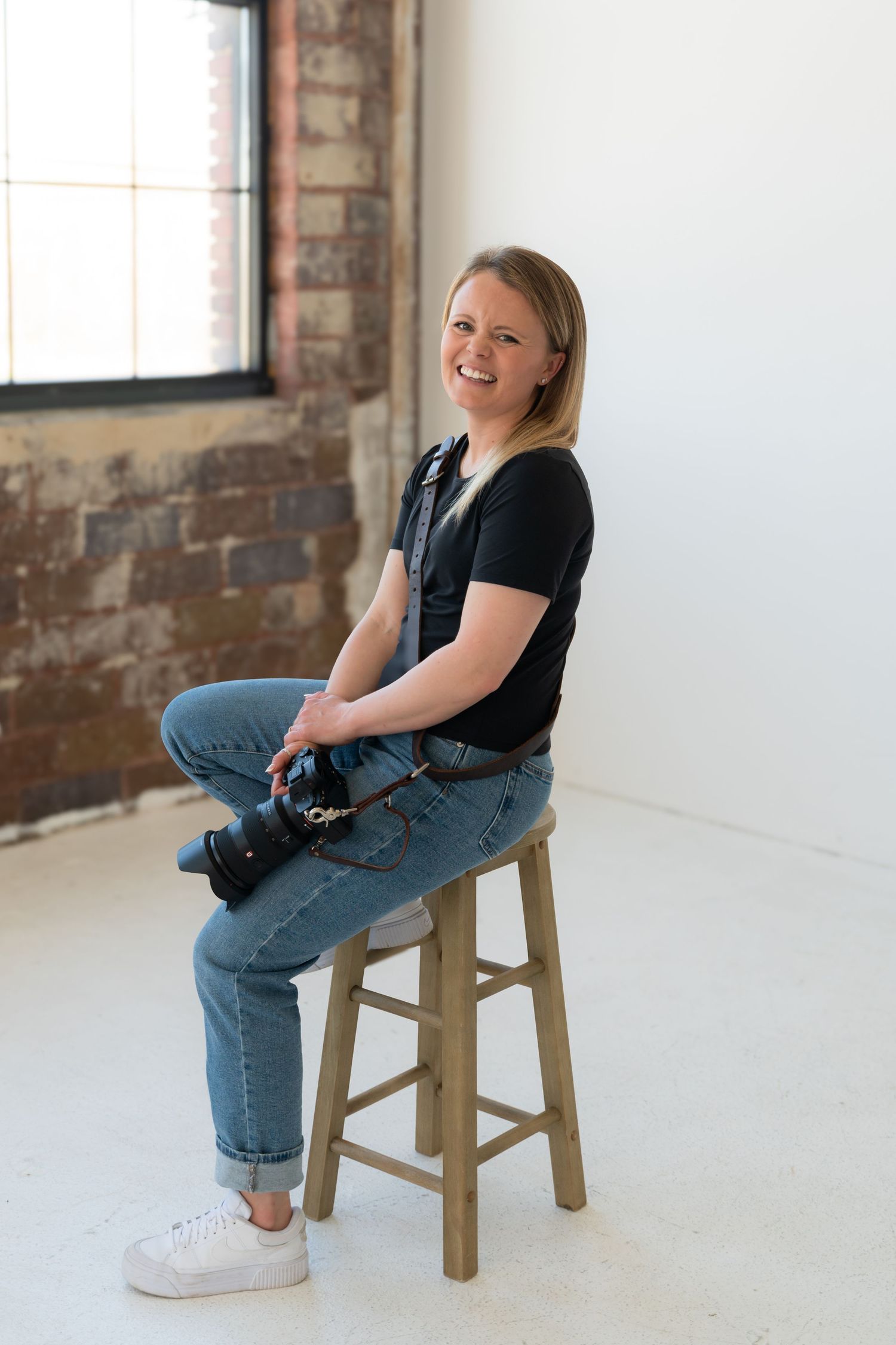 Casual portrait session showing seated person in black top and jeans on wooden stool against white and brick wall backdrop.