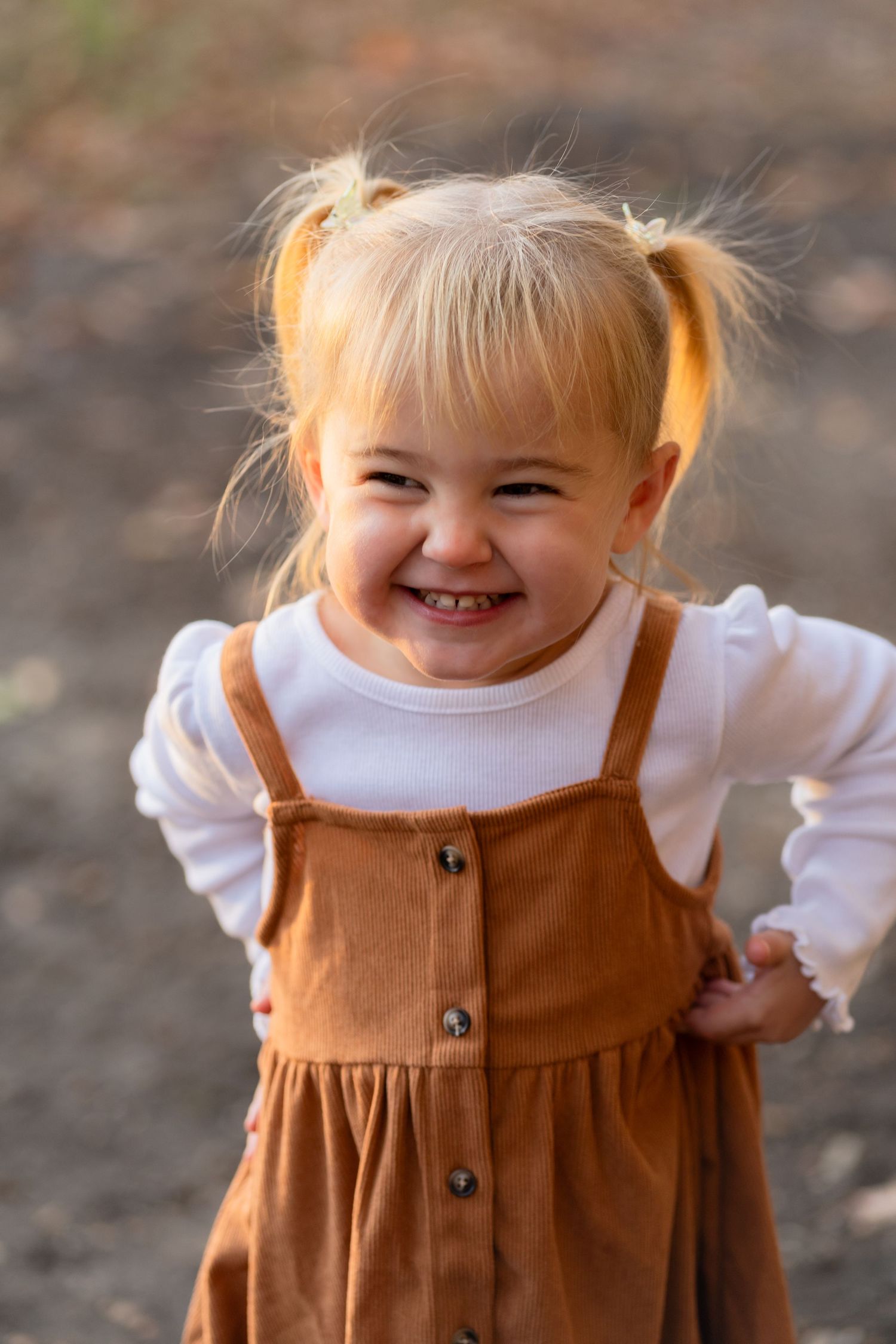 Smiling child in brown corduroy overalls and white shirt with blonde pigtails standing outdoors.