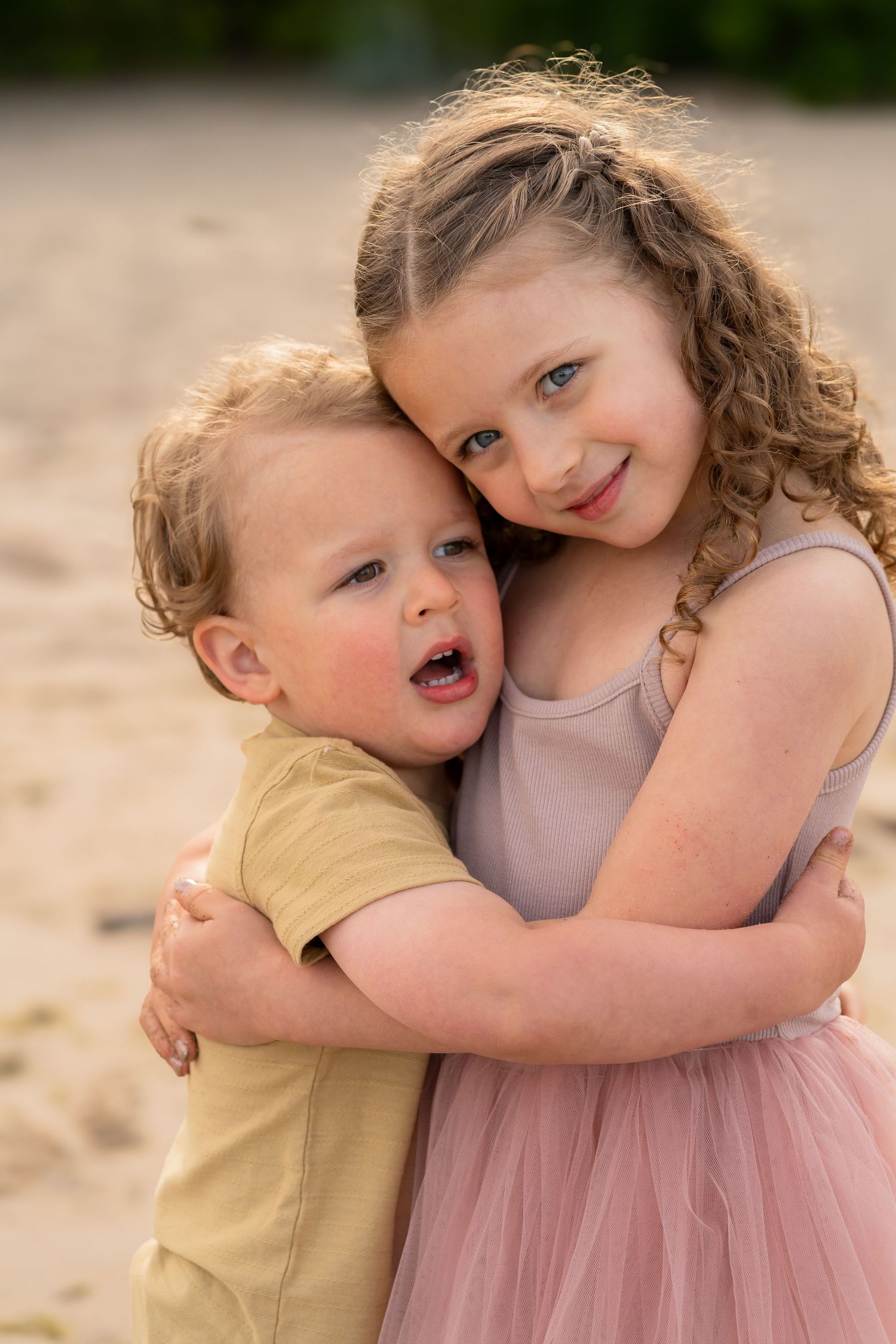Two young children embrace and smile while standing on sand, one wearing yellow and the other in a pink tulle dress.