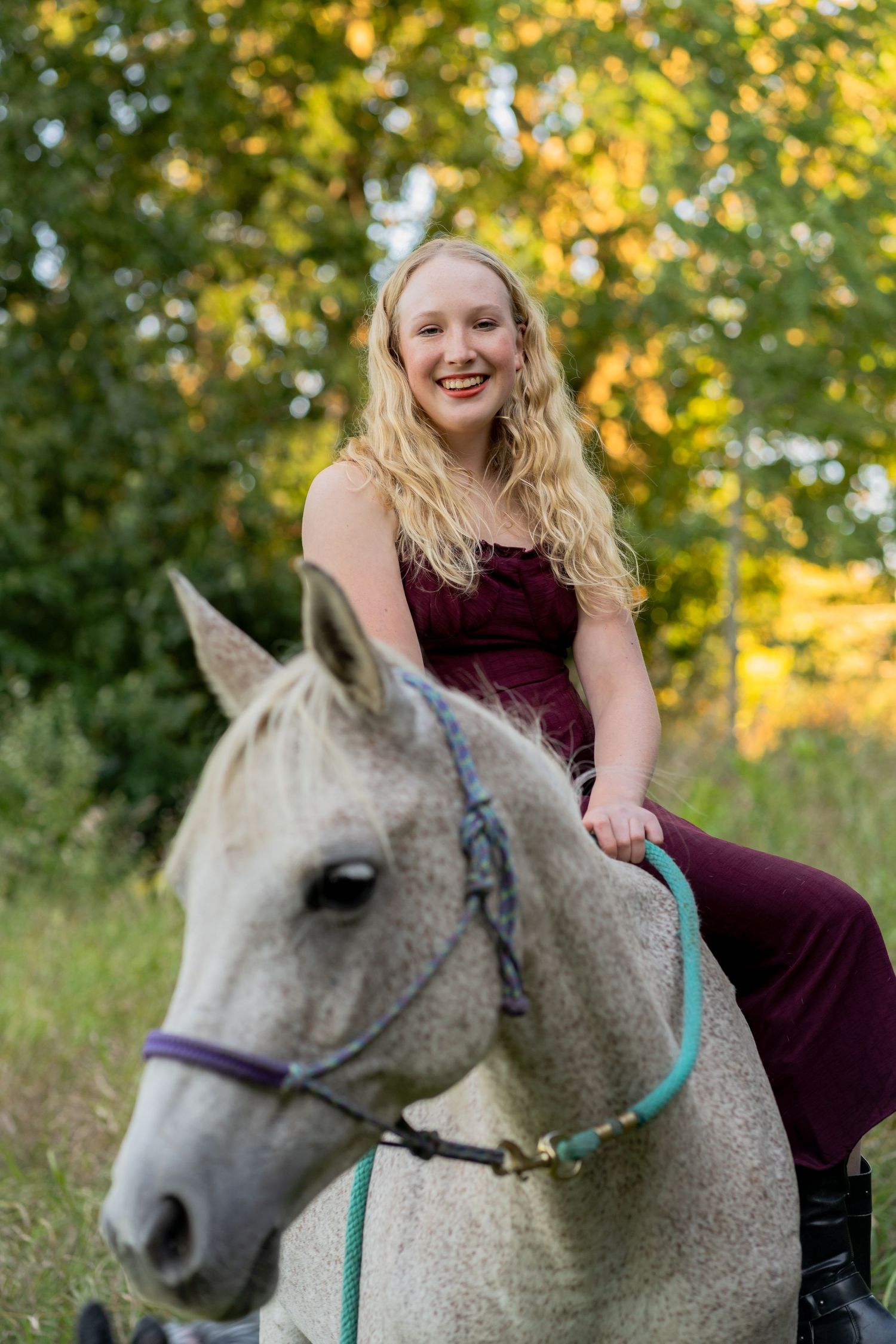 A person in a burgundy dress sits atop a dappled grey horse in a sunny outdoor setting with autumn foliage.