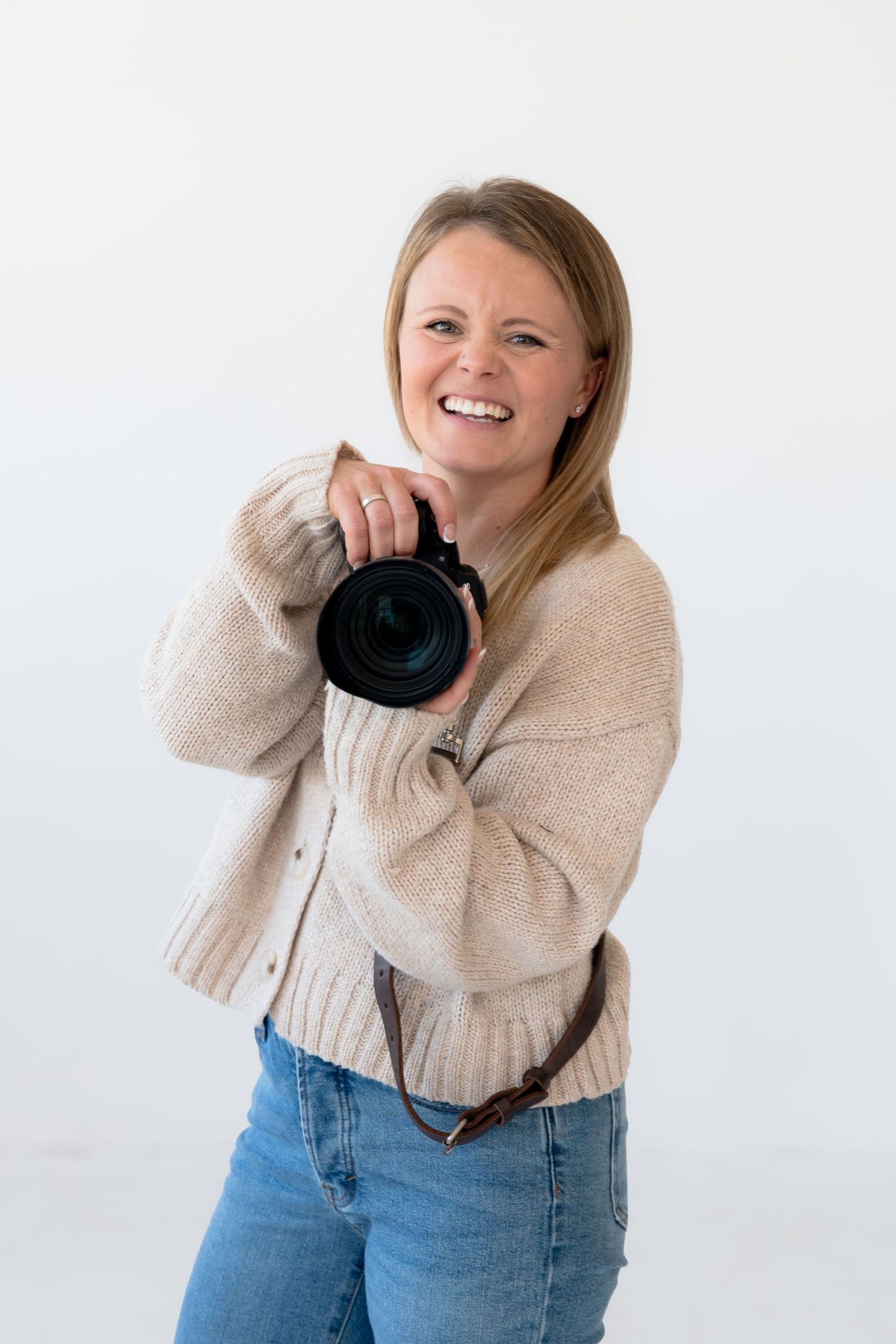 A photographer in a beige sweater and jeans holds a professional camera while posing against a white background.