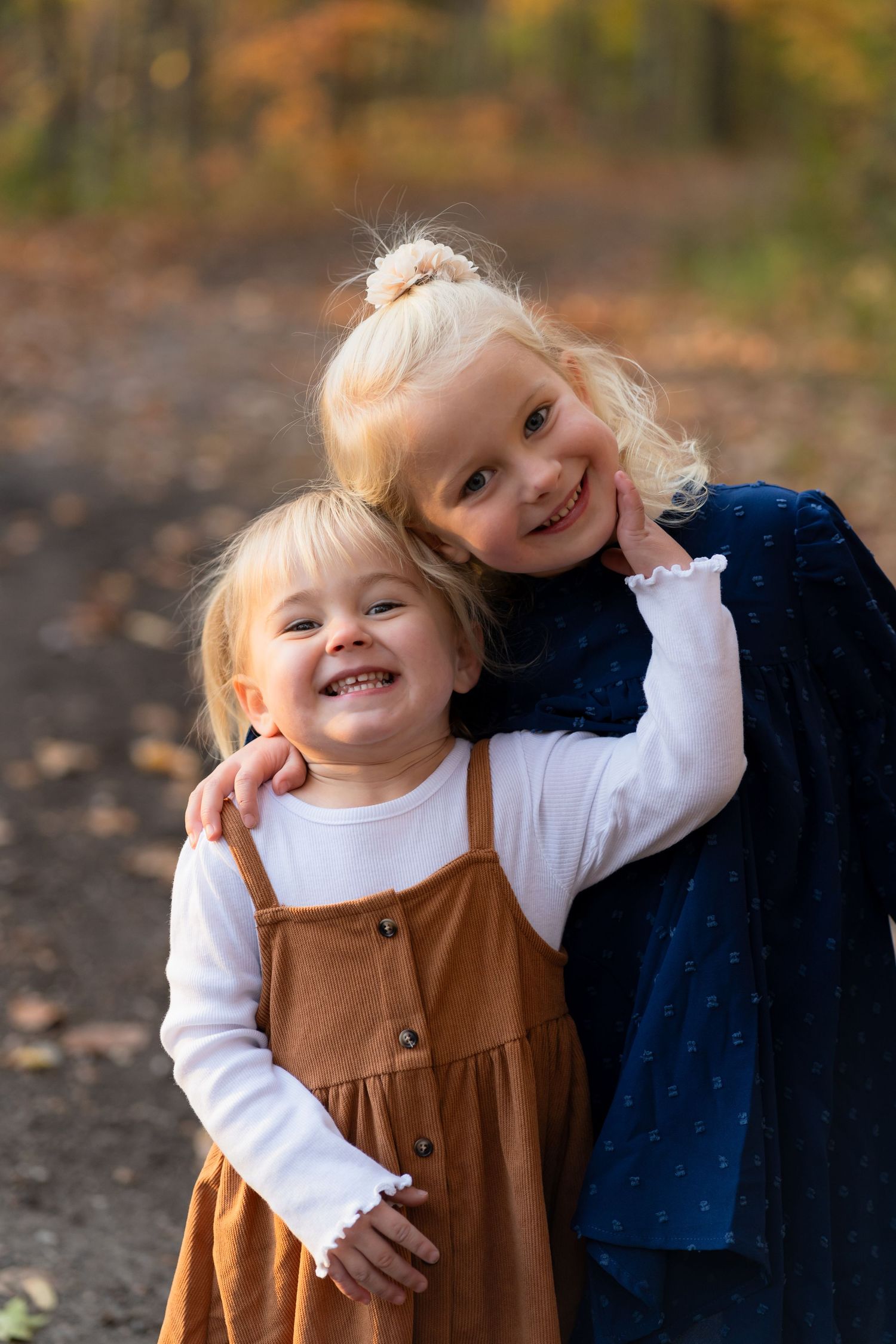 Two children in fall dresses embrace and smile outdoors on an autumn day with fallen leaves in the background.