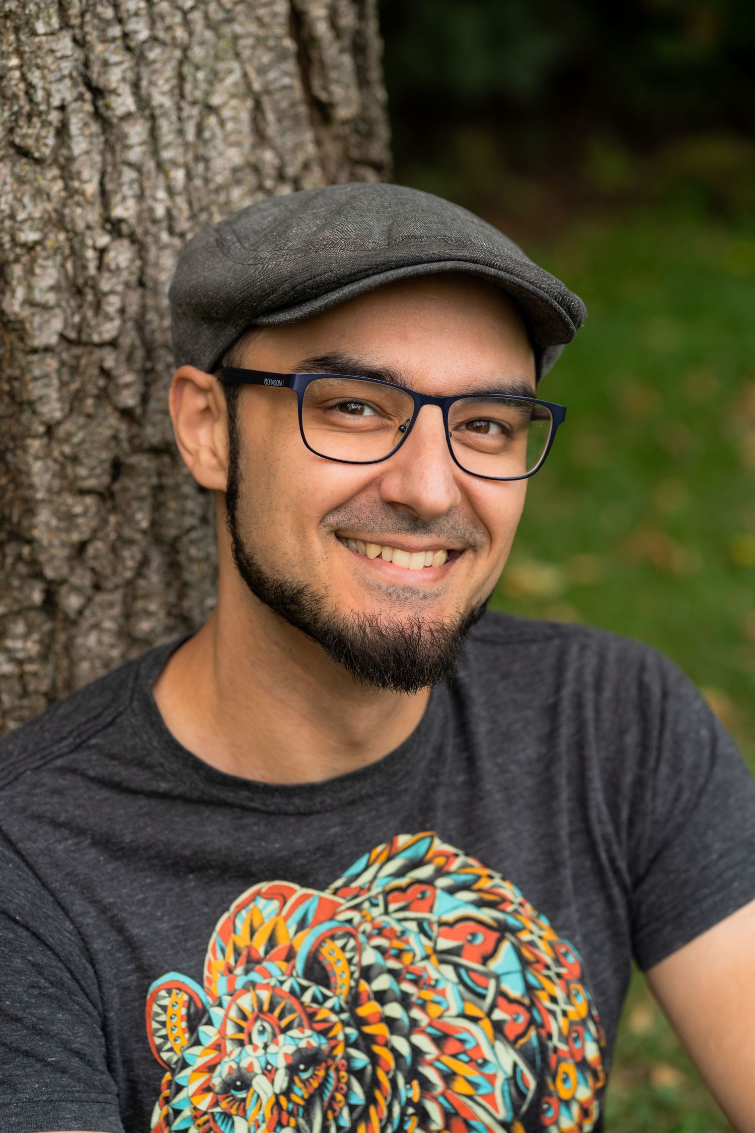 Outdoor portrait of someone in a gray flat cap and glasses wearing a colorful patterned t-shirt while leaning against a tree.
