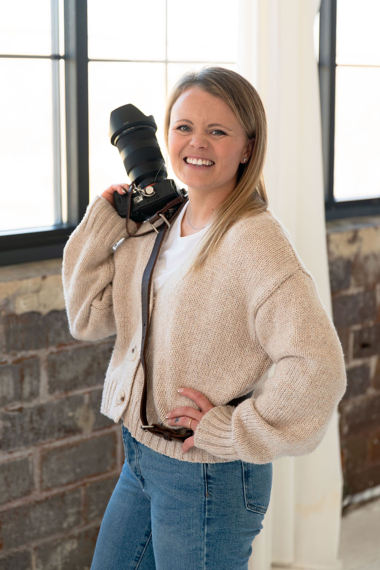 A photographer holding a professional DSLR camera stands in a cozy studio space wearing a beige cardigan and jeans.