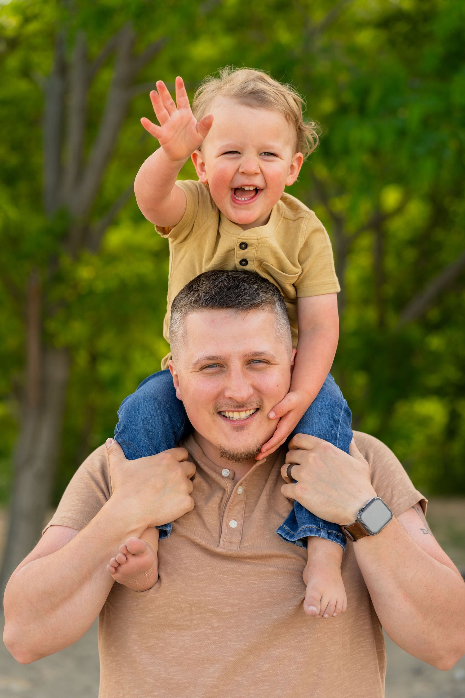 A joyful moment outdoors as a child rides on shoulders, both smiling with green foliage in the background.
