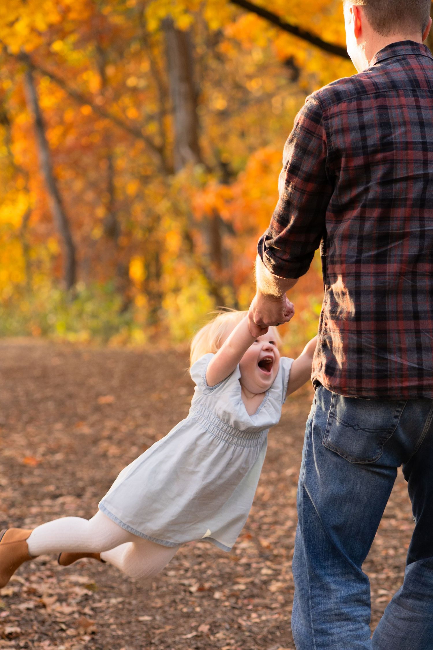 A person in a plaid shirt swings a small child wearing a white dress among autumn foliage in a park.