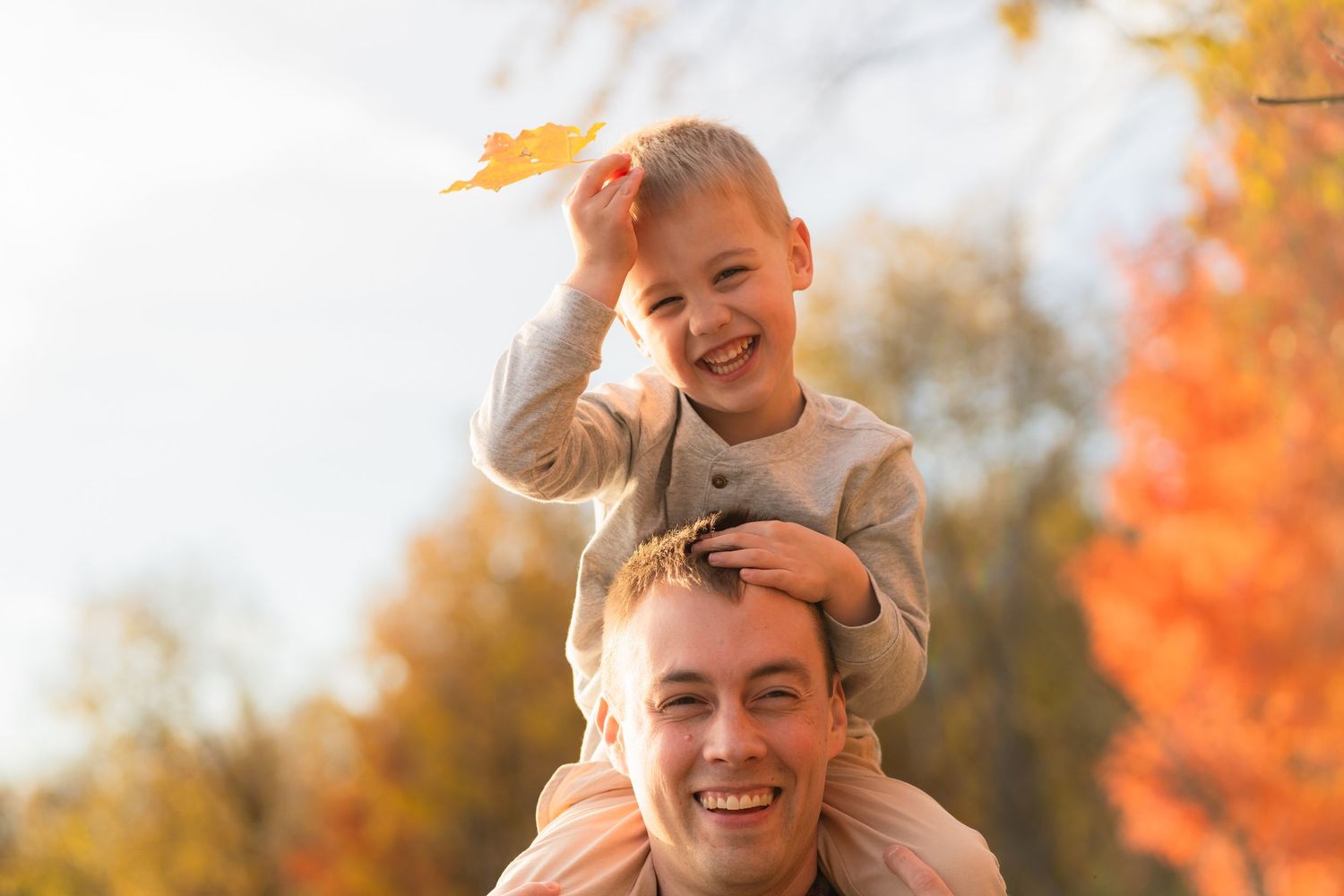 A young child perched on an adult's shoulders outdoors amid vibrant autumn foliage on a sunny day.