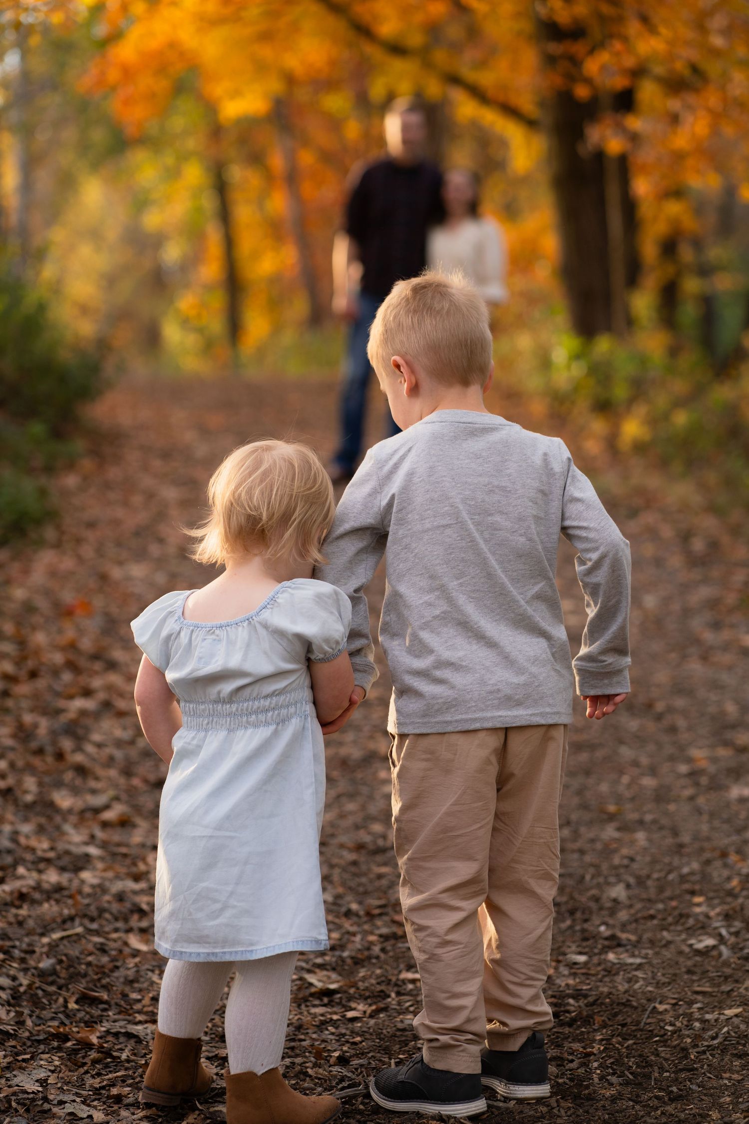 Two children walk along a wooded path covered in fallen leaves during autumn with golden foliage in the background.
