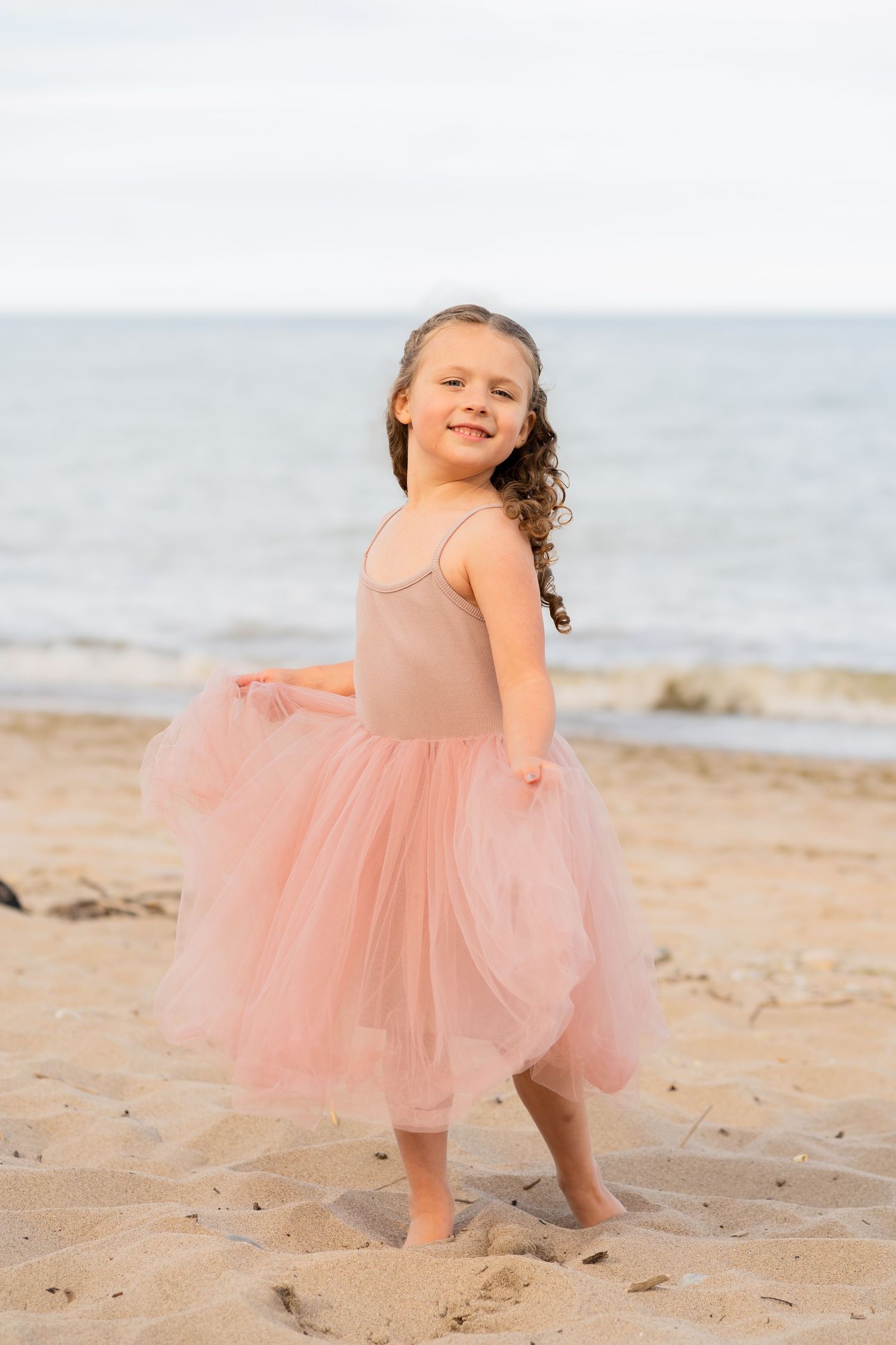 Young dancer in pink tulle dress twirls on beach sand with ocean waves in background.