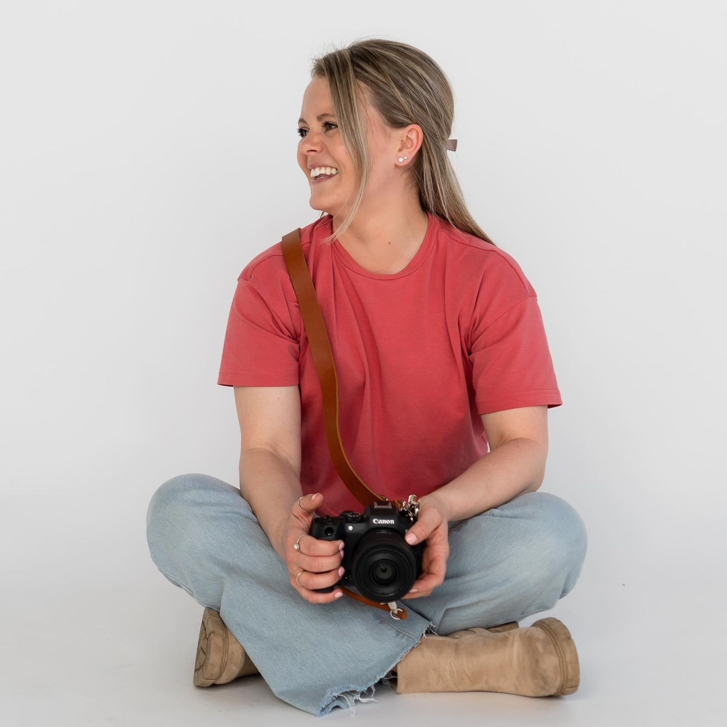 Person in red t-shirt and jeans sits cross-legged on white background while laughing and holding a camera.