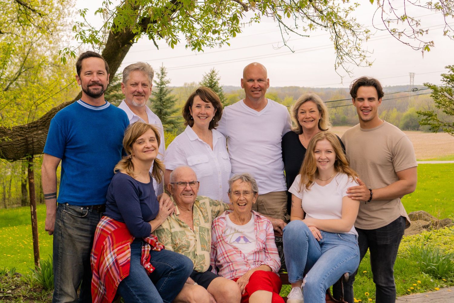 A group of people gather for a casual outdoor family portrait in a grassy field with trees in the background.