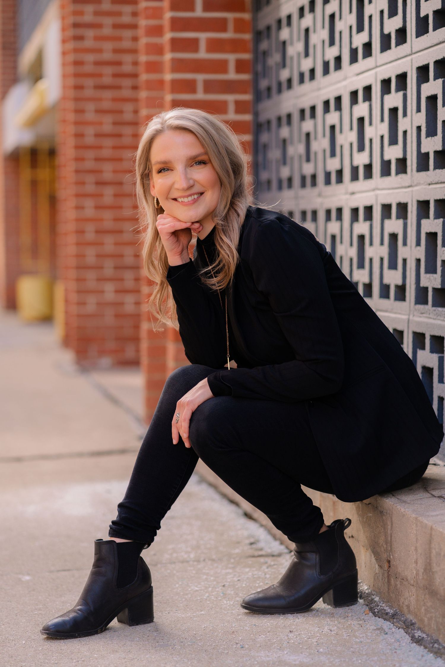 A woman in an all-black outfit with ankle boots crouches and smiles near a decorative brick wall.