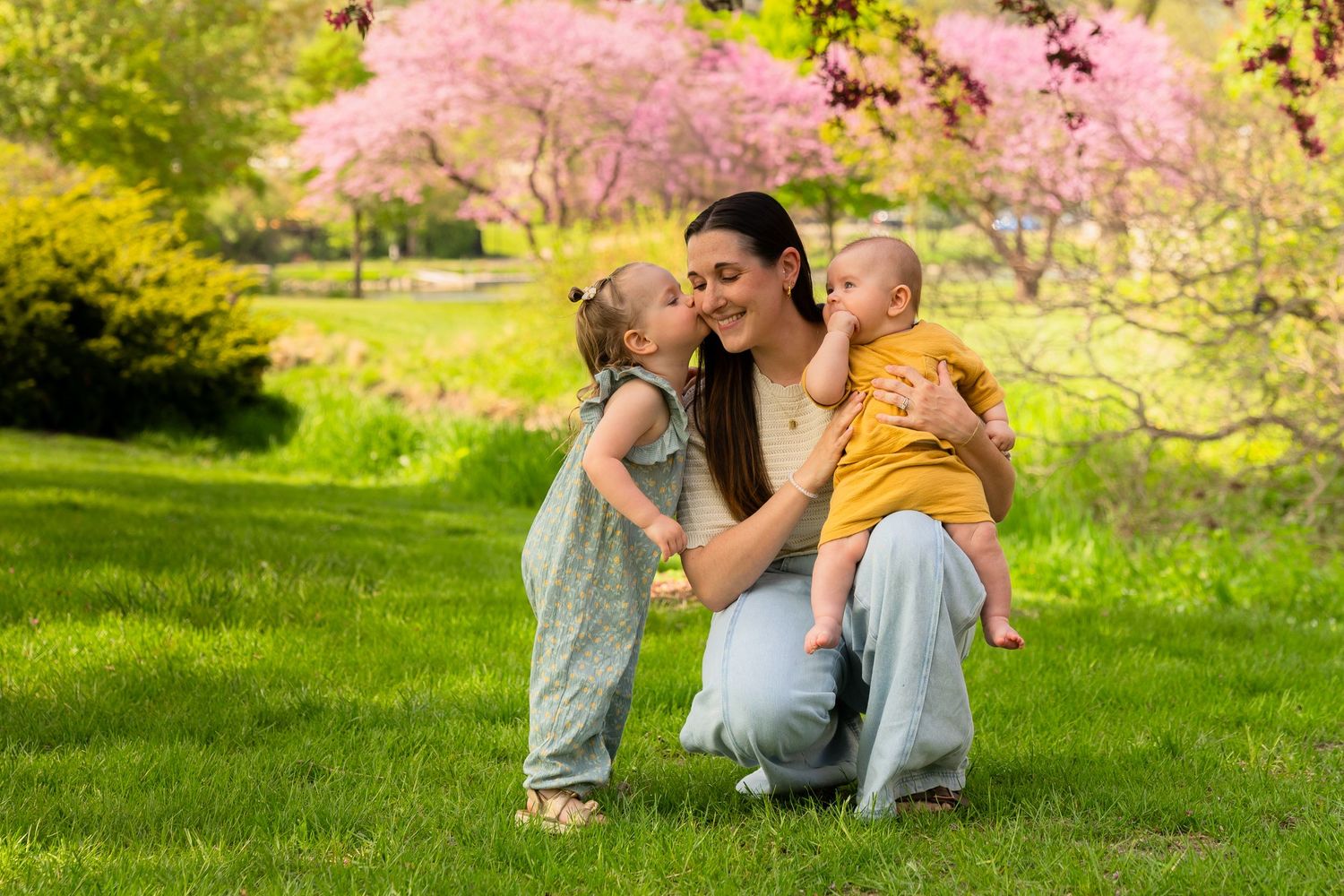 A tender family moment shared outdoors during spring with blooming cherry blossoms and lush green grass in the background.