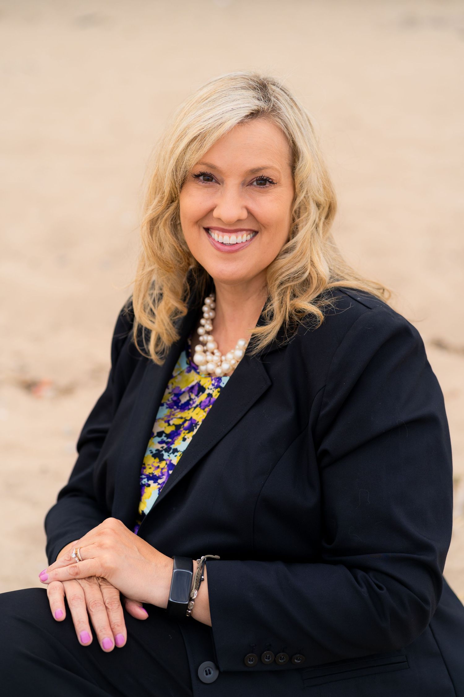 Professional headshot of a blonde woman in a black blazer and colorful statement necklace smiling warmly against a neutral background.