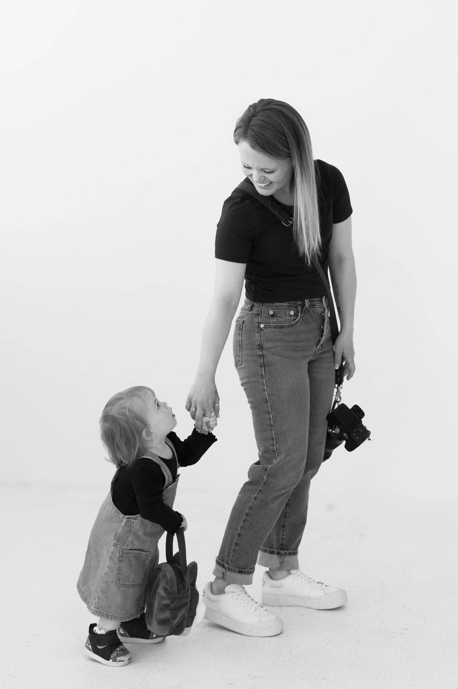 Black and white portrait of photographer with camera standing beside small child holding matching camera bag against white wall.