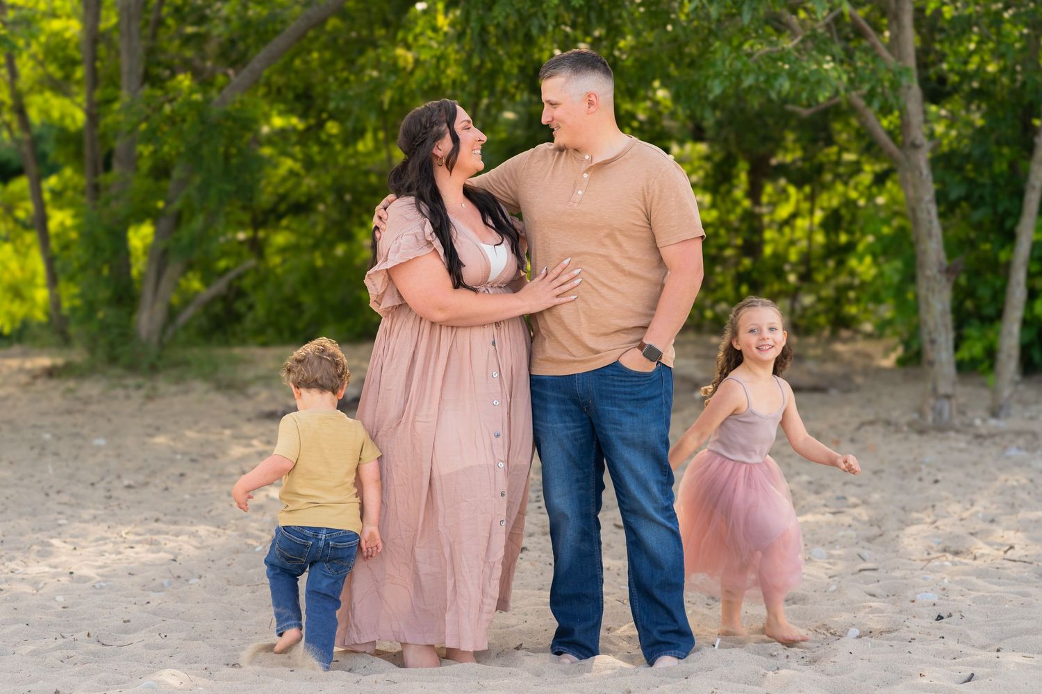 A family of four stands together on a sandy beach path surrounded by green trees in warm evening light.
