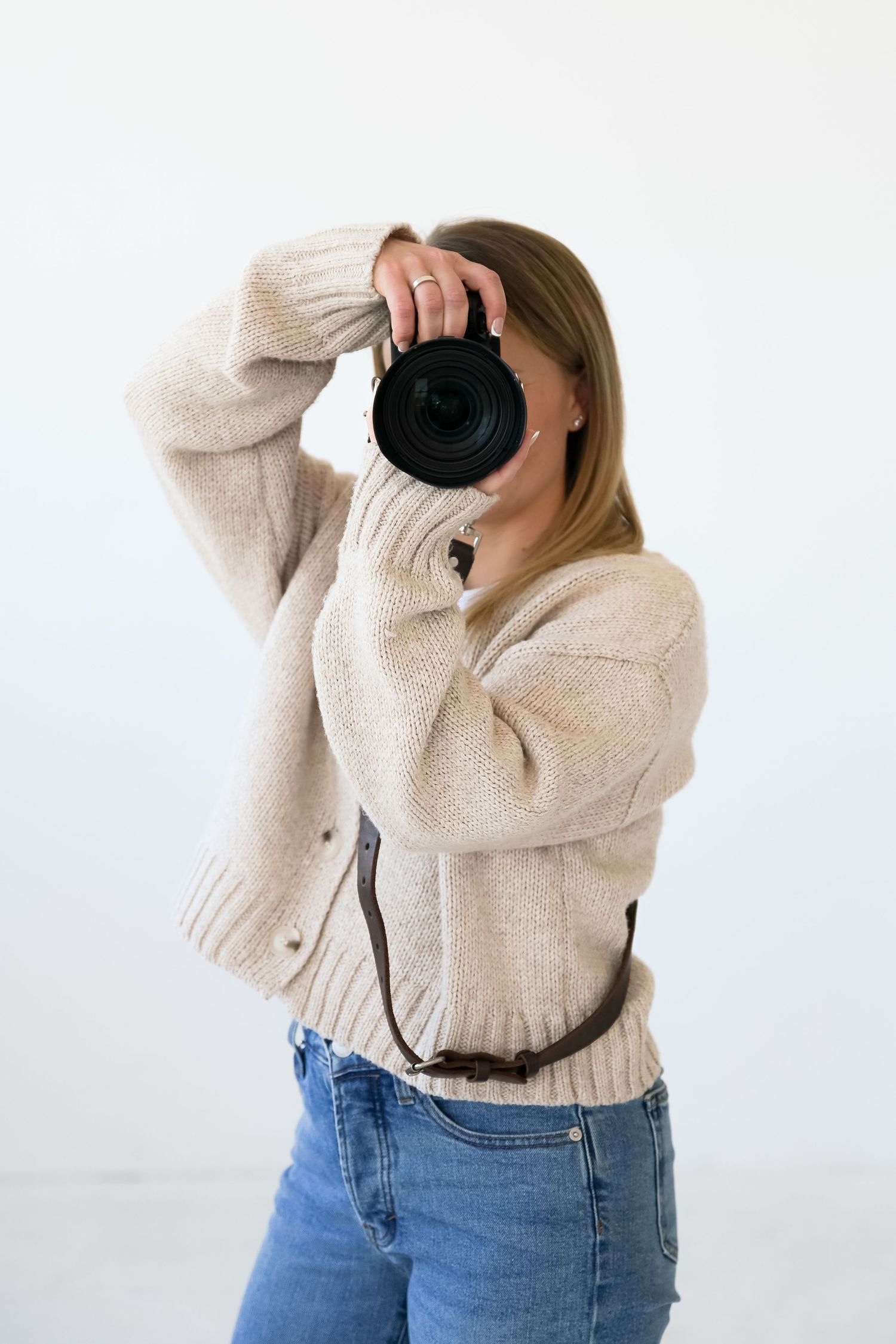 Photographer wearing cozy beige sweater and jeans holds up professional camera against white background.
