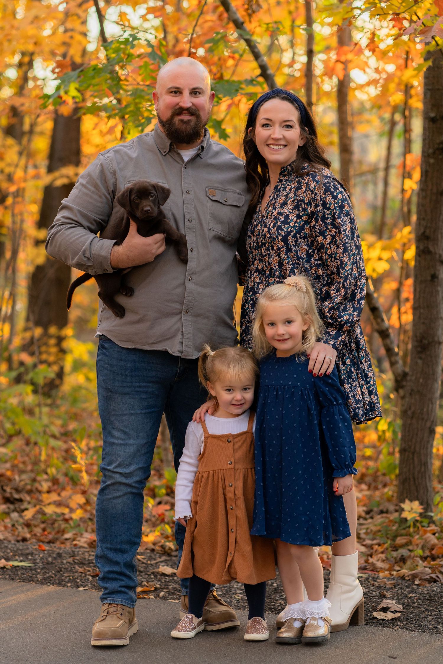 A family poses together on a path surrounded by vibrant fall foliage while holding a small brown puppy.