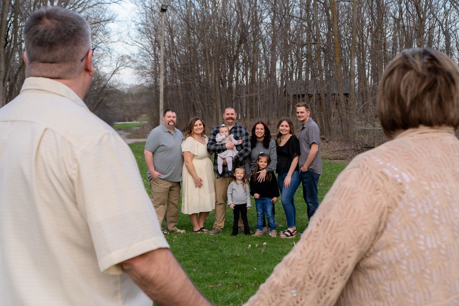 A multi-generational family gathers outdoors on a grassy area with bare trees in the background.